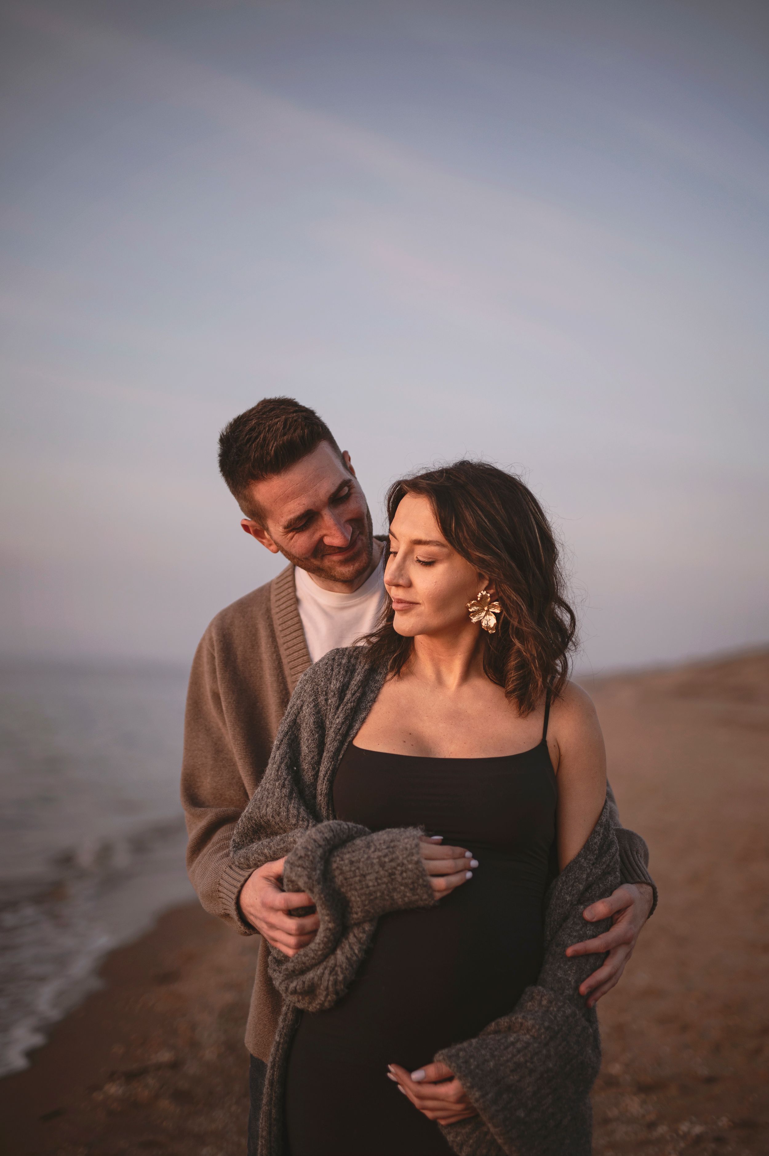 A romantic couple shares an intimate moment at sunset on a beach, with one wearing a cozy sweater and the other in black.