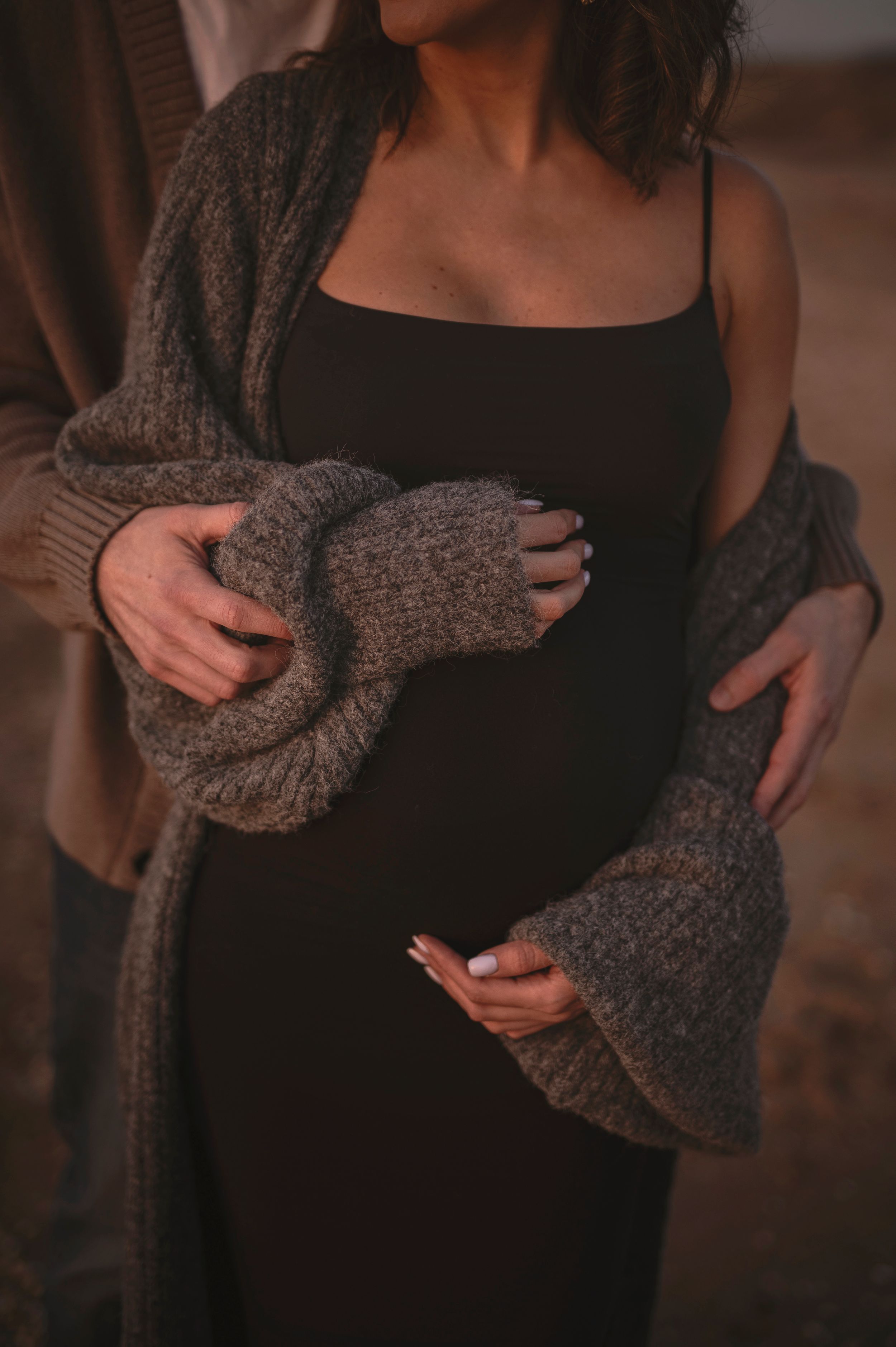 Someone wearing a cozy gray knit cardigan over a black slip dress in warm moody lighting.