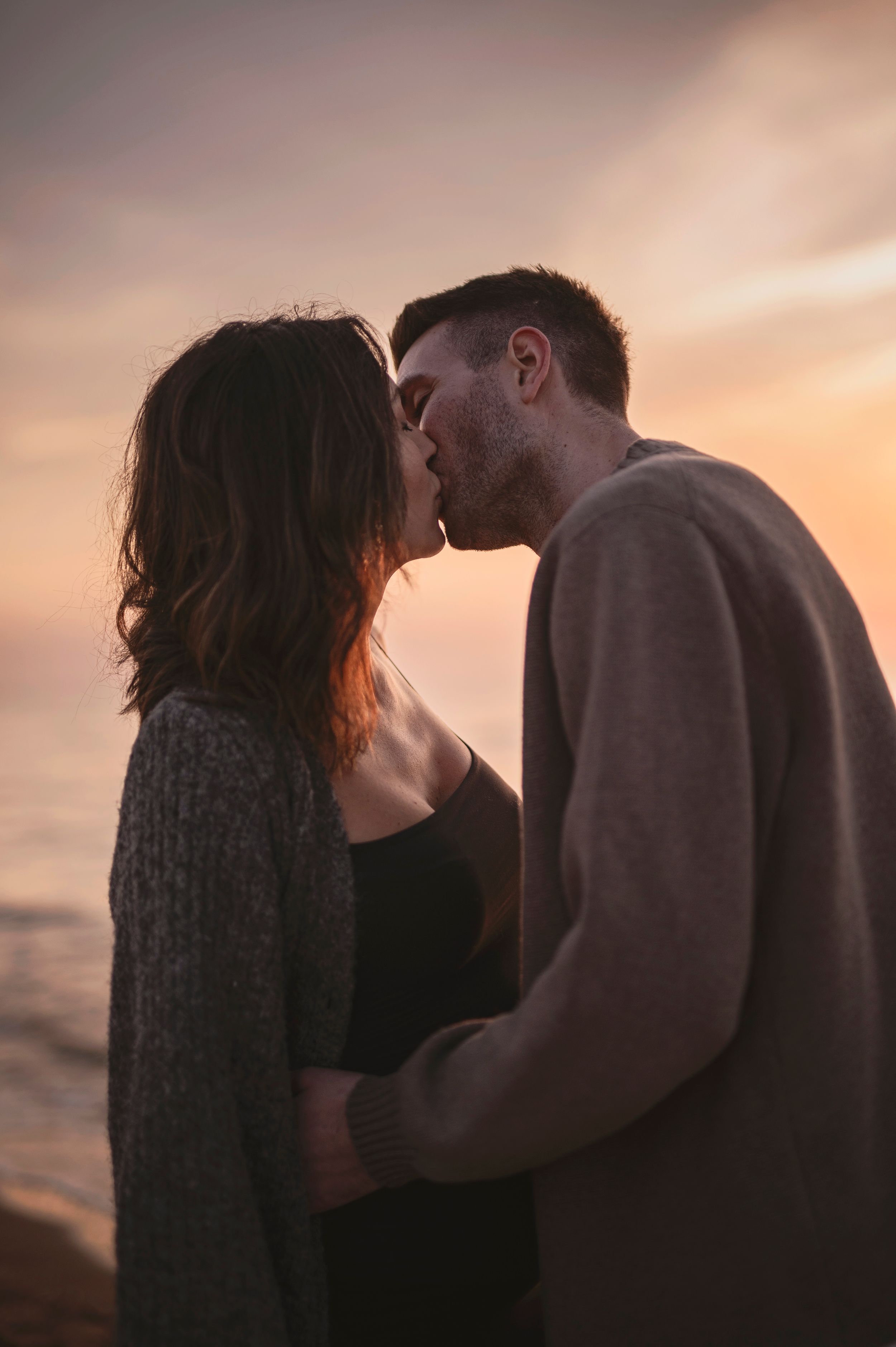A romantic couple shares a tender kiss during sunset by the ocean, silhouetted against a warm glowing sky.