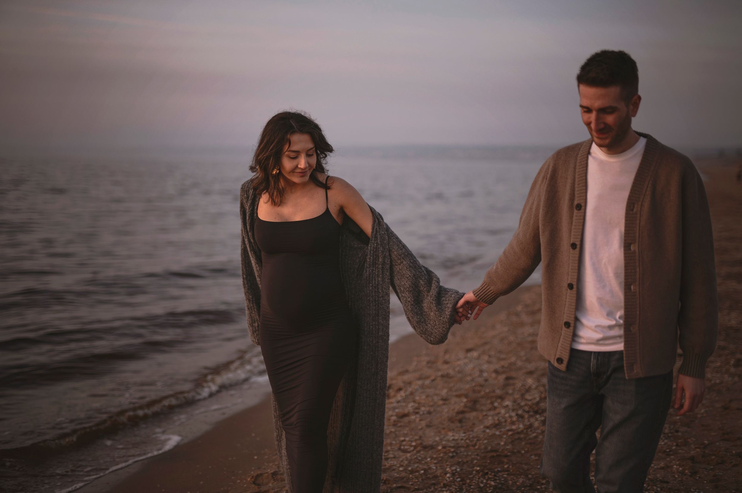 A couple walks hand in hand along a moody beach shoreline at dusk.