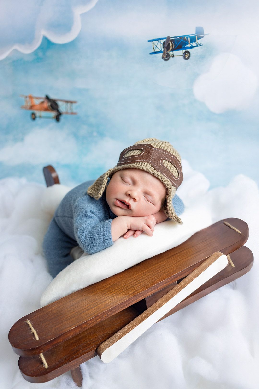 A sleeping baby in a winter hat lies on clouds with vintage toy planes and wooden skis in a dreamy aviation themed photo.