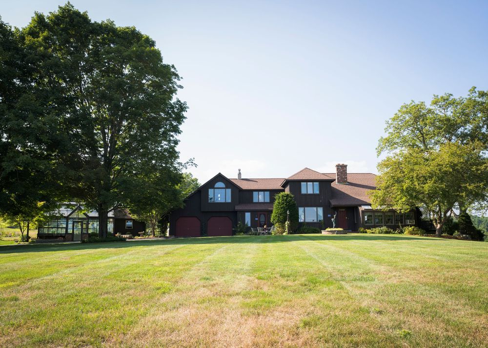 Large red New England style home with dormers and green lawn surrounded by mature trees on sunny summer day.