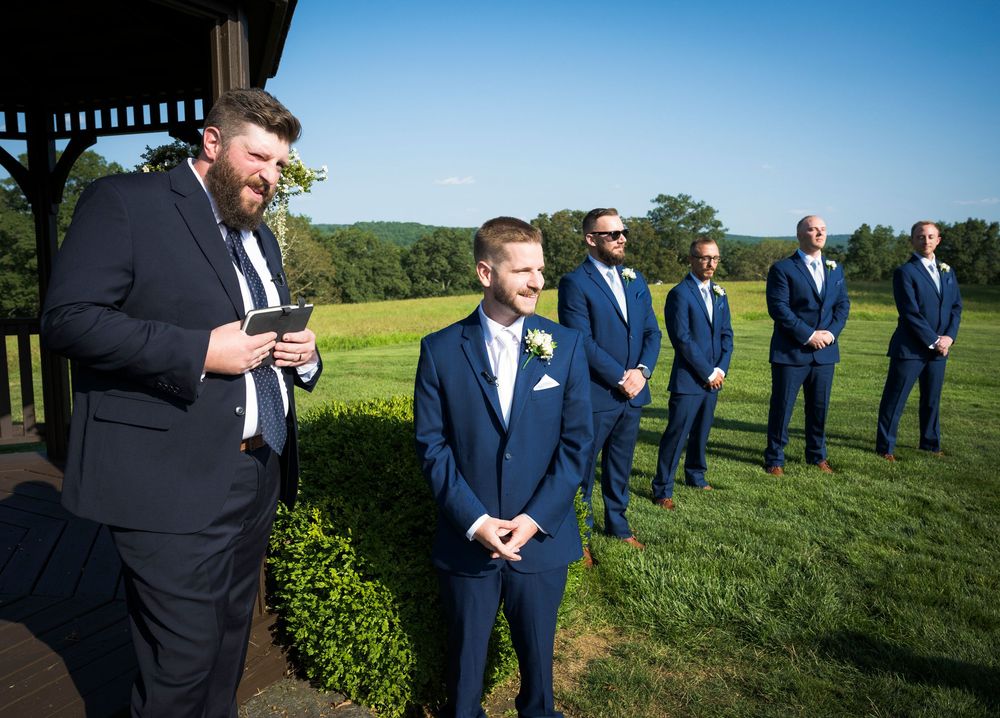 Wedding party lined up on grassy hill against bright blue sky wearing dark suits.