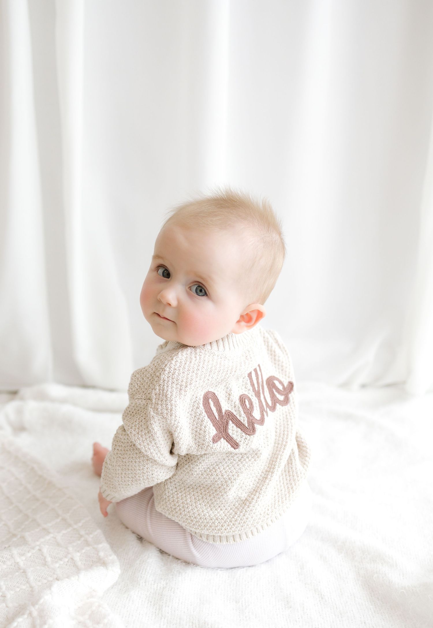 Baby wearing a cream knit sweater with hello script, sitting on white blanket.