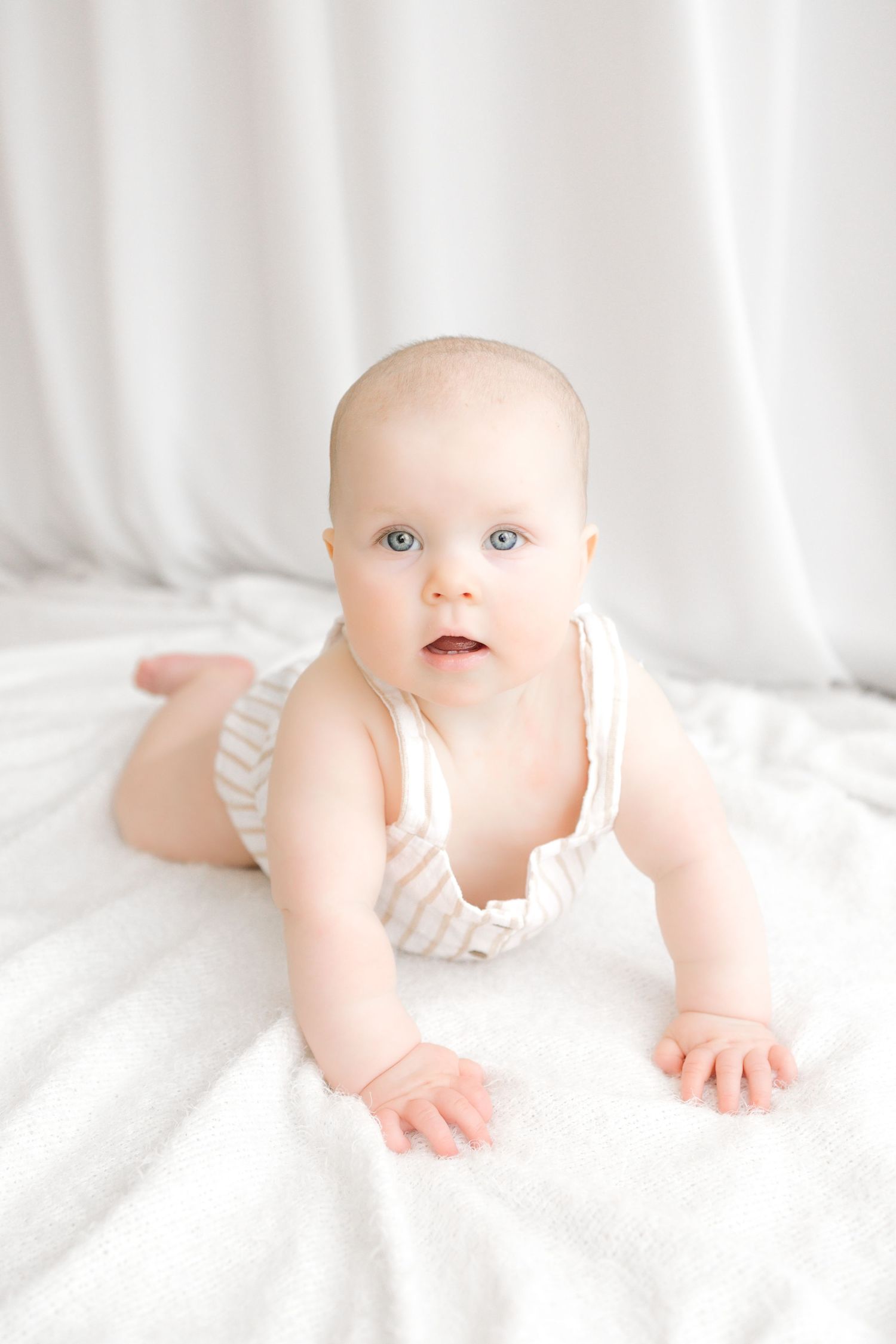 A cute baby with blue eyes in a striped romper doing tummy time on a white blanket.