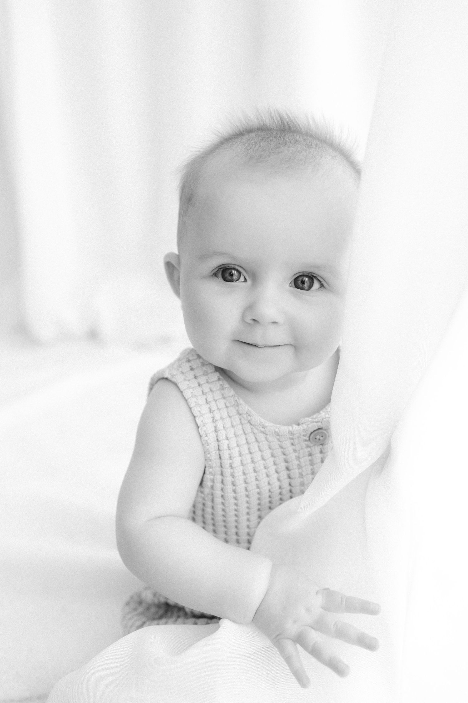 Black and white portrait of an adorable smiling baby in a knit romper peeking around a white curtain.