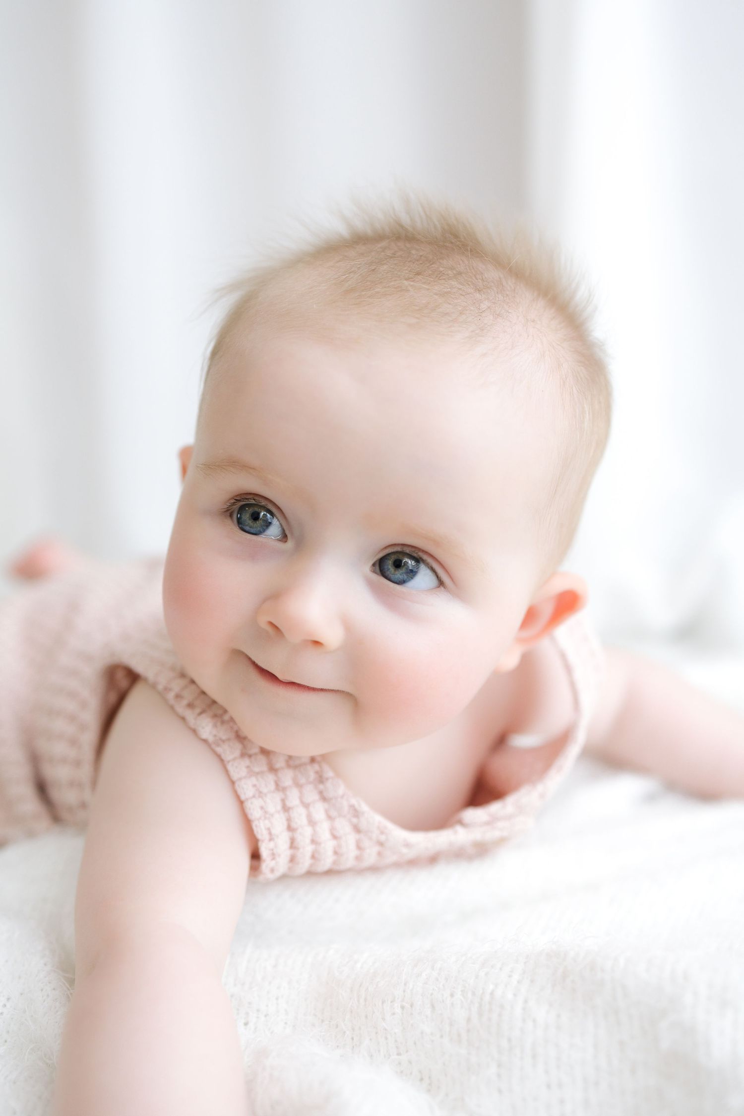 Adorable baby with blue eyes in pink knit outfit lying on white blanket, looking up with a sweet smile.