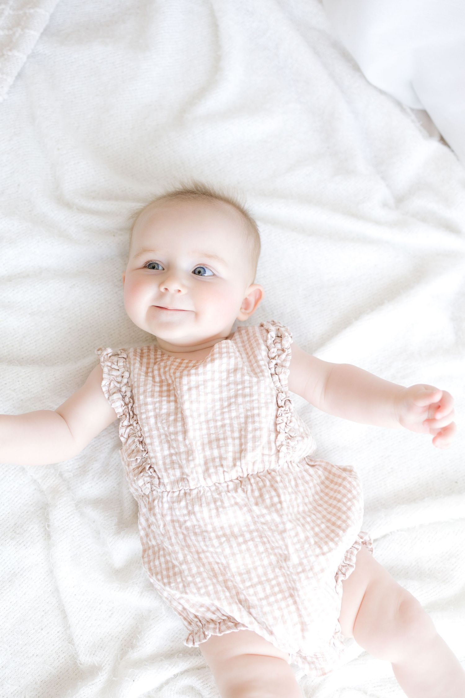 Smiling baby girl in a pink gingham ruffle romper lying on a white blanket, looking up with bright blue eyes.