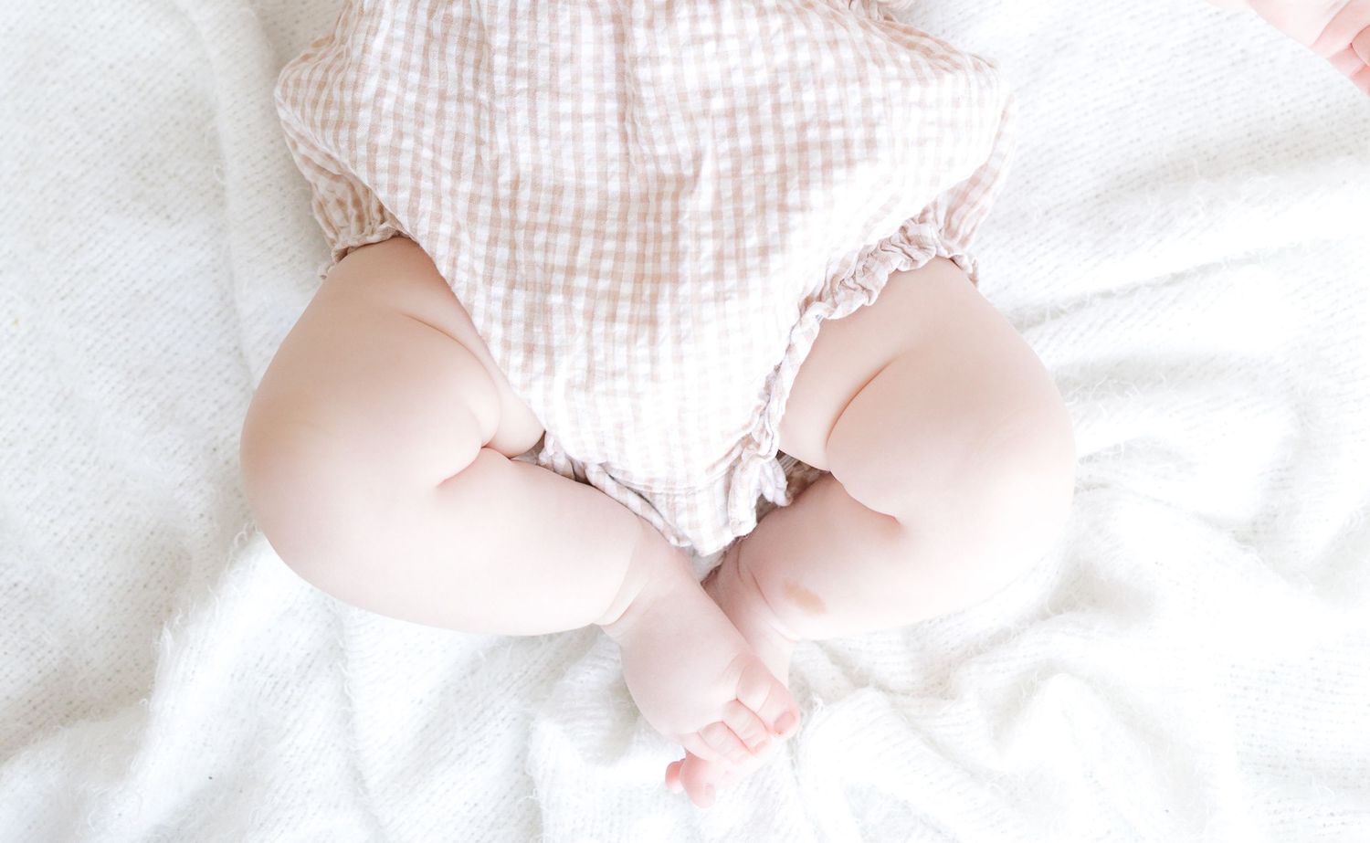 Baby in pink gingham romper lying on white blanket, chubby legs and tiny feet visible from above.