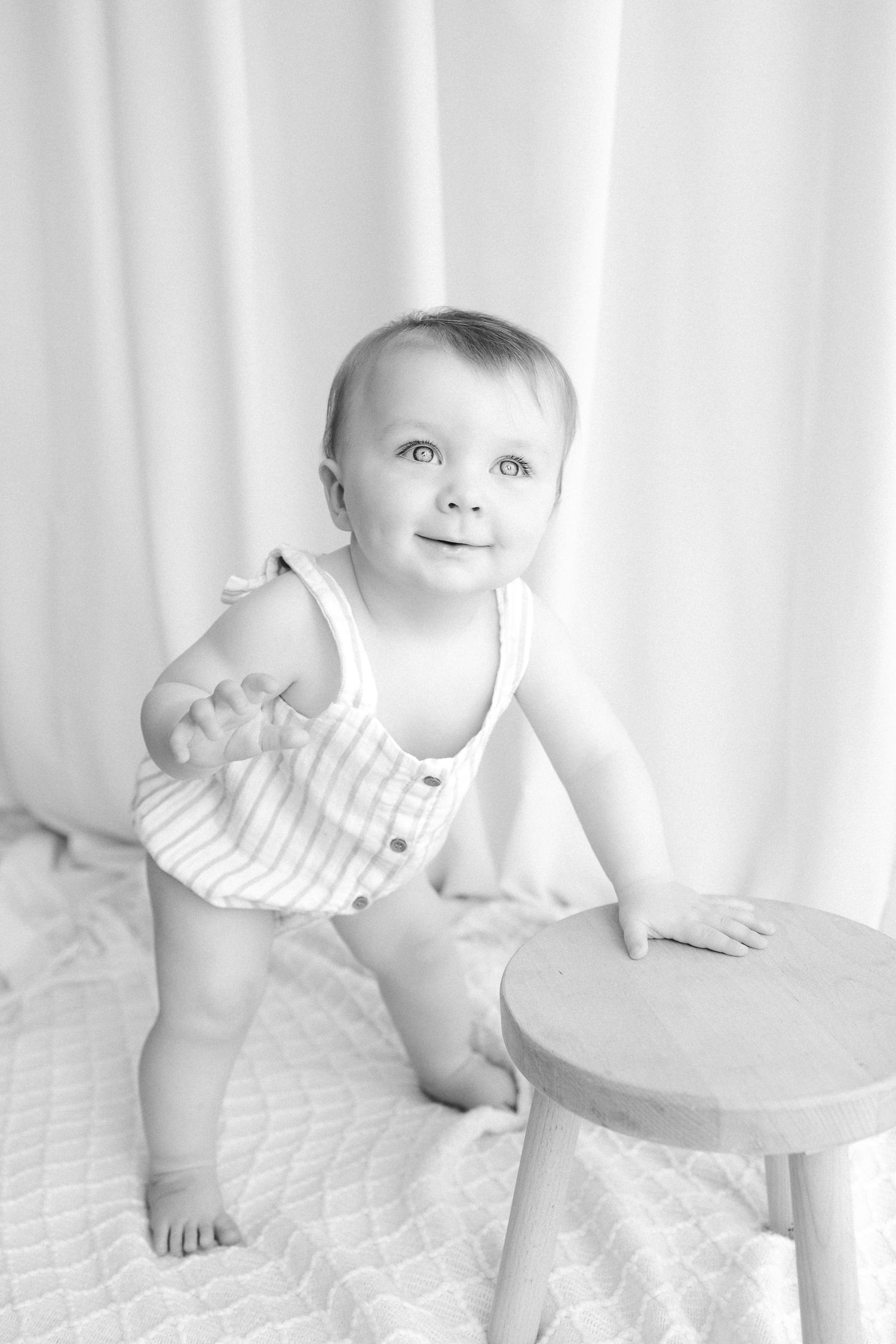 Black and white photo of an adorable baby in a striped romper standing next to a small wooden stool.