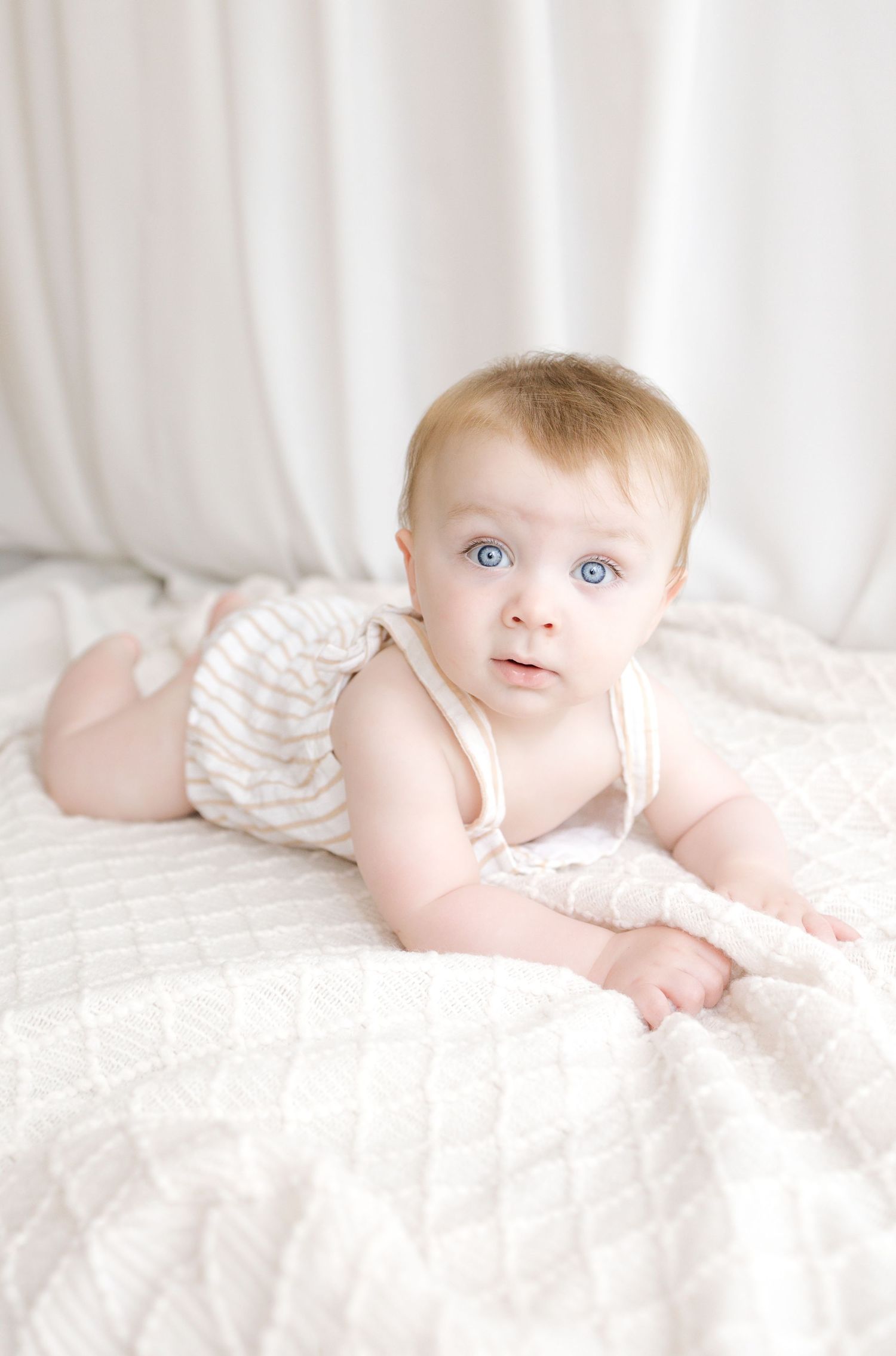 Blue-eyed baby in striped romper doing tummy time on white quilted blanket with soft white background.