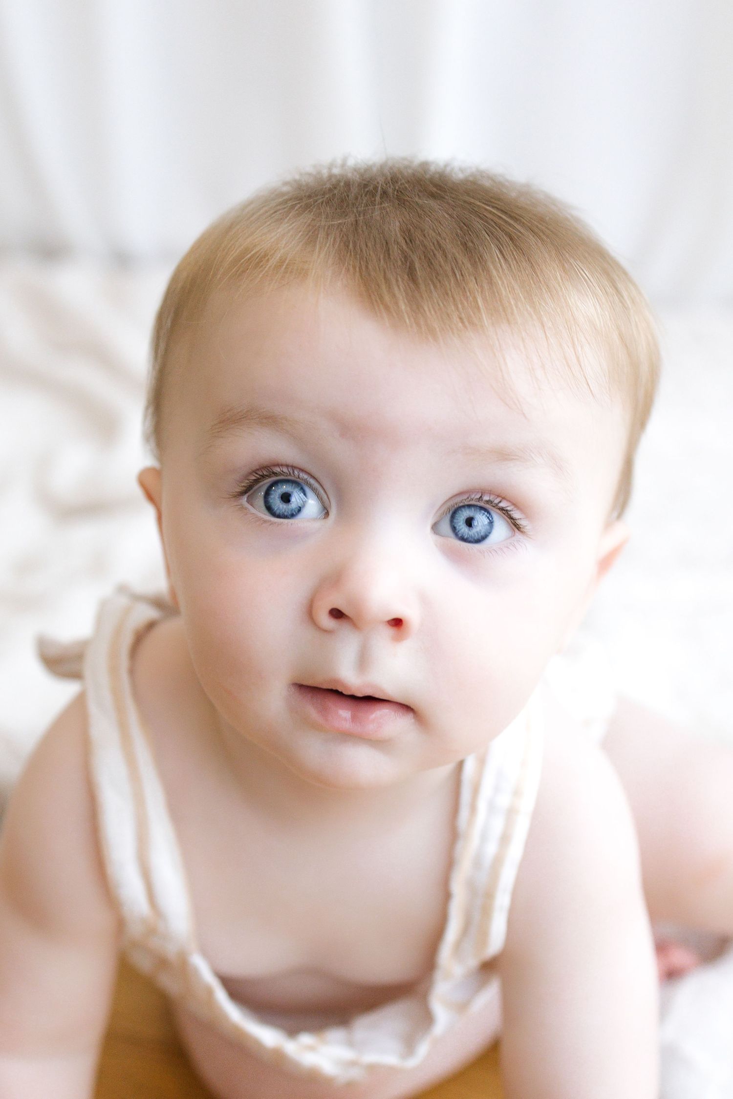 Adorable baby with big blue eyes and light hair looks curiously at camera while lying on white bedding.