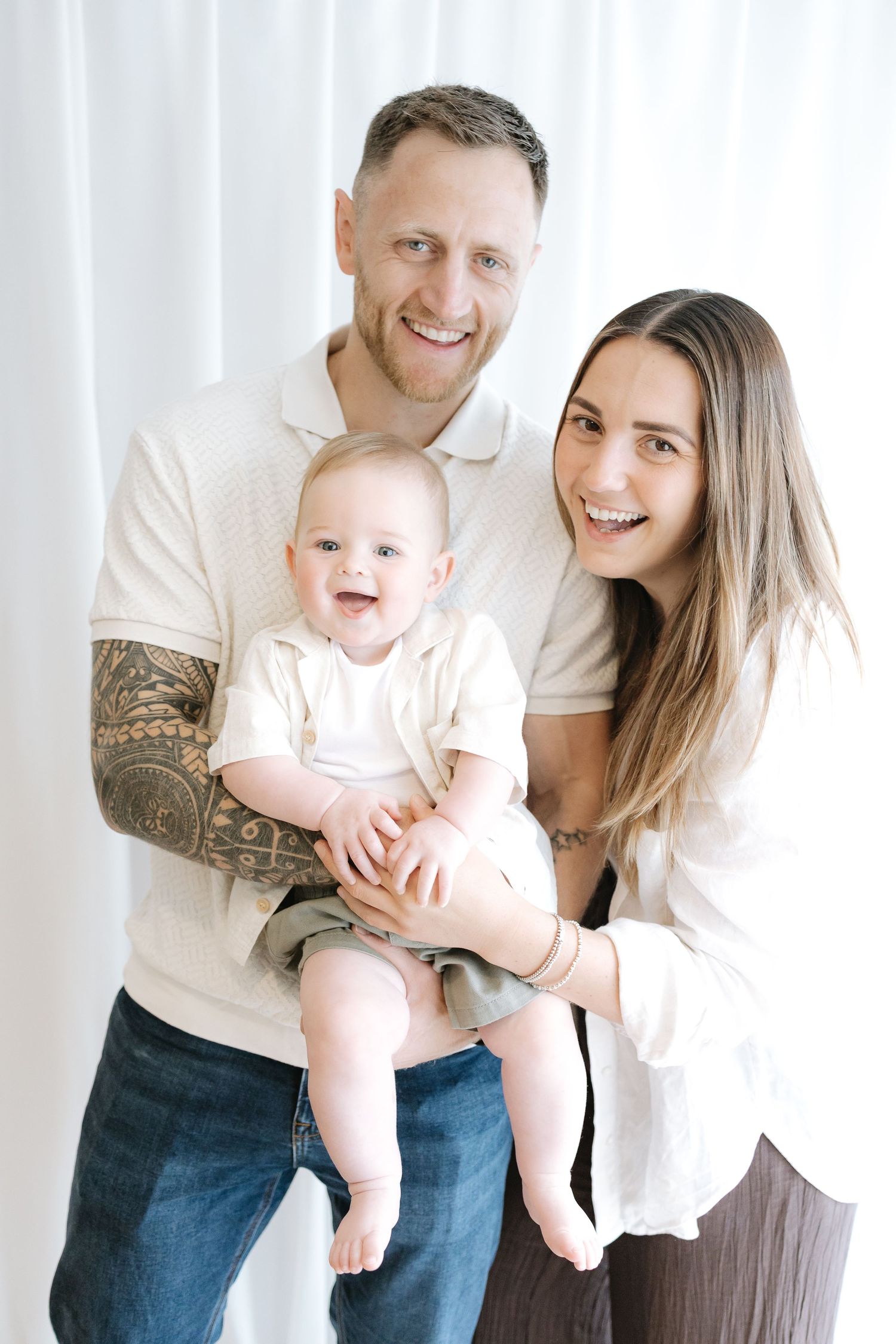 Smiling couple holding a happy baby in a bright white studio, man with tattoos wearing a beige polo shirt.