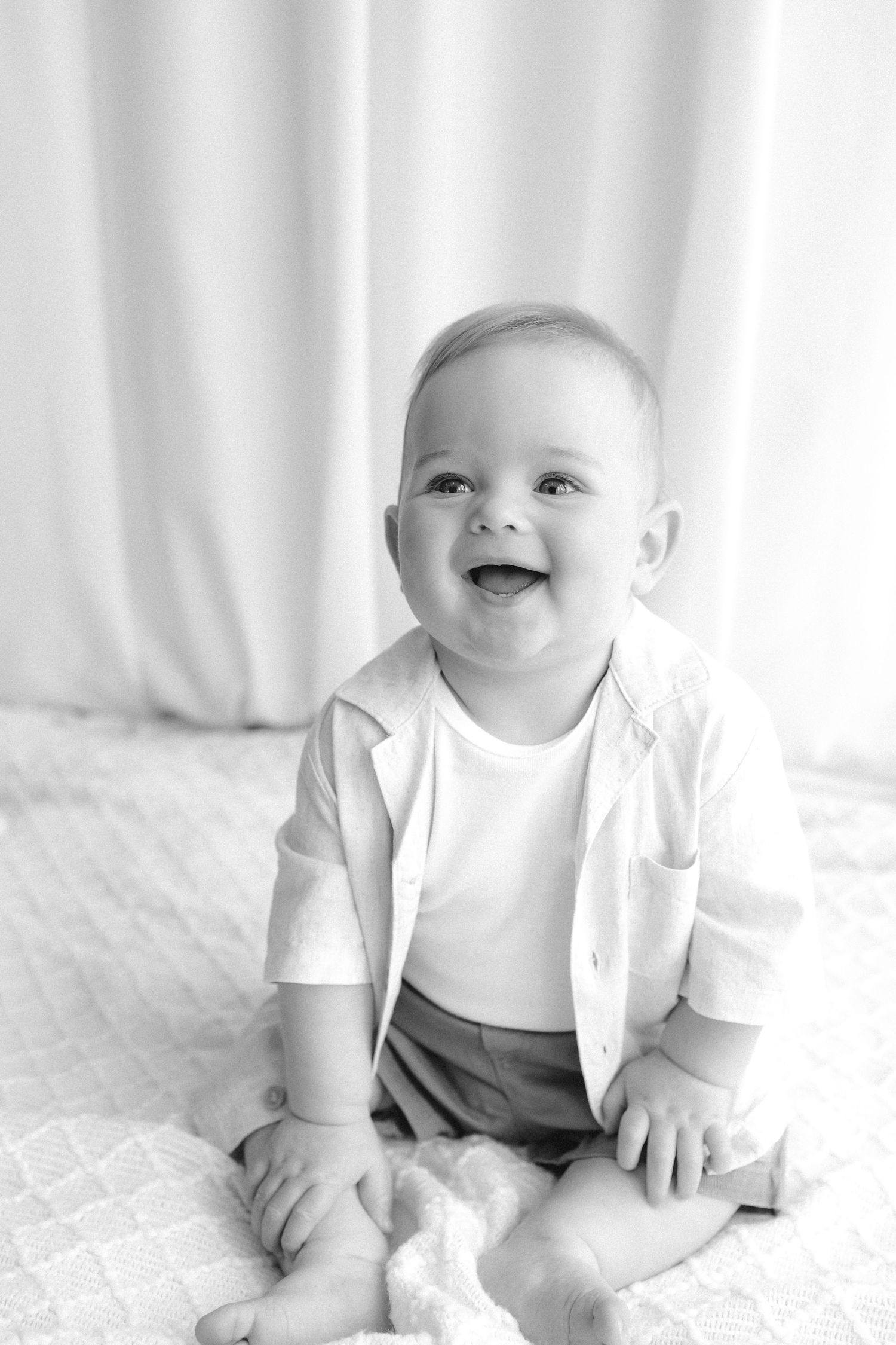Black and white photo of a happy laughing baby boy sitting up, wearing a white shirt and shorts.