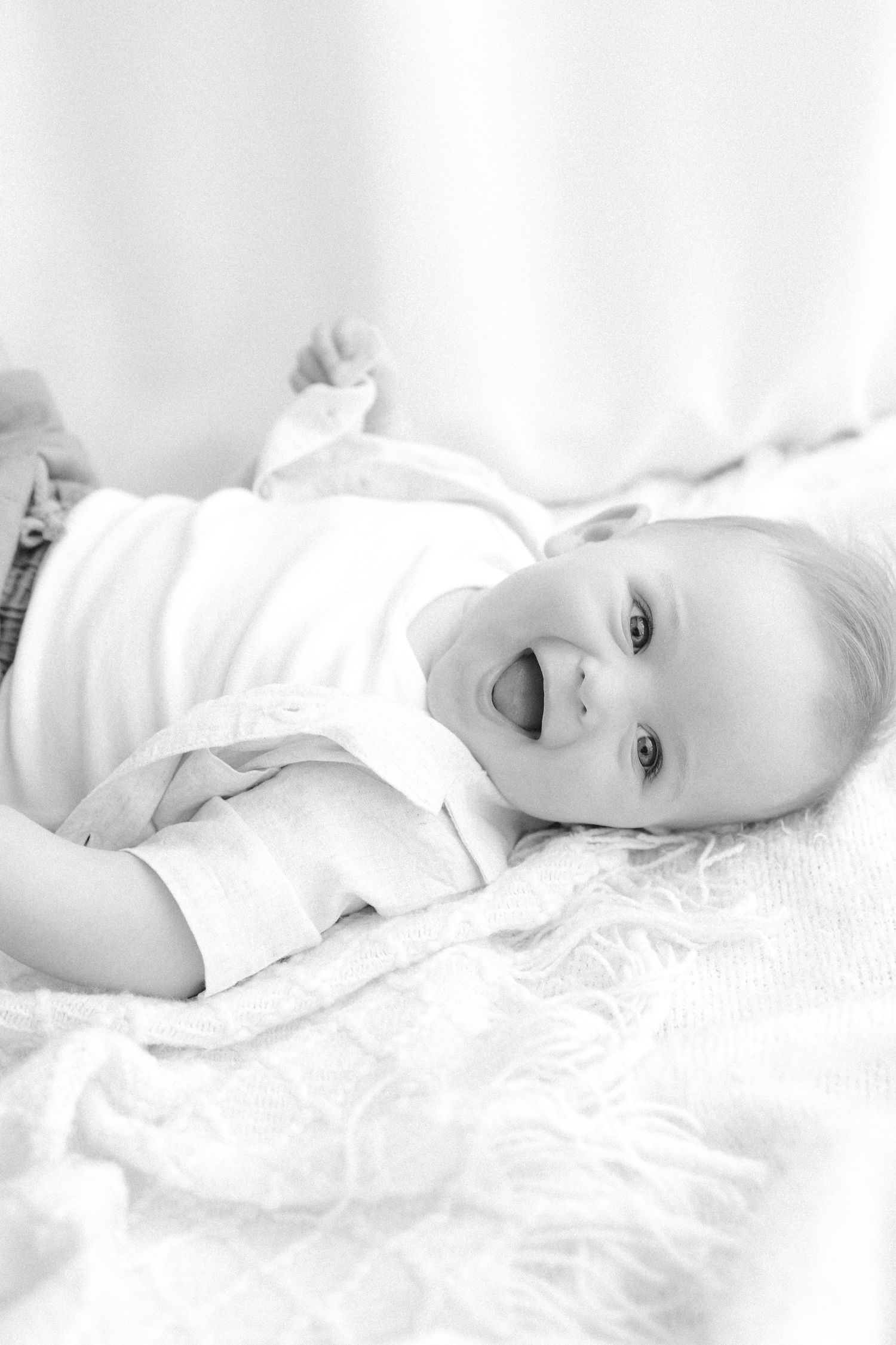 Black and white photo of a happy laughing baby lying on a white blanket, looking up with bright eyes and an open smile.