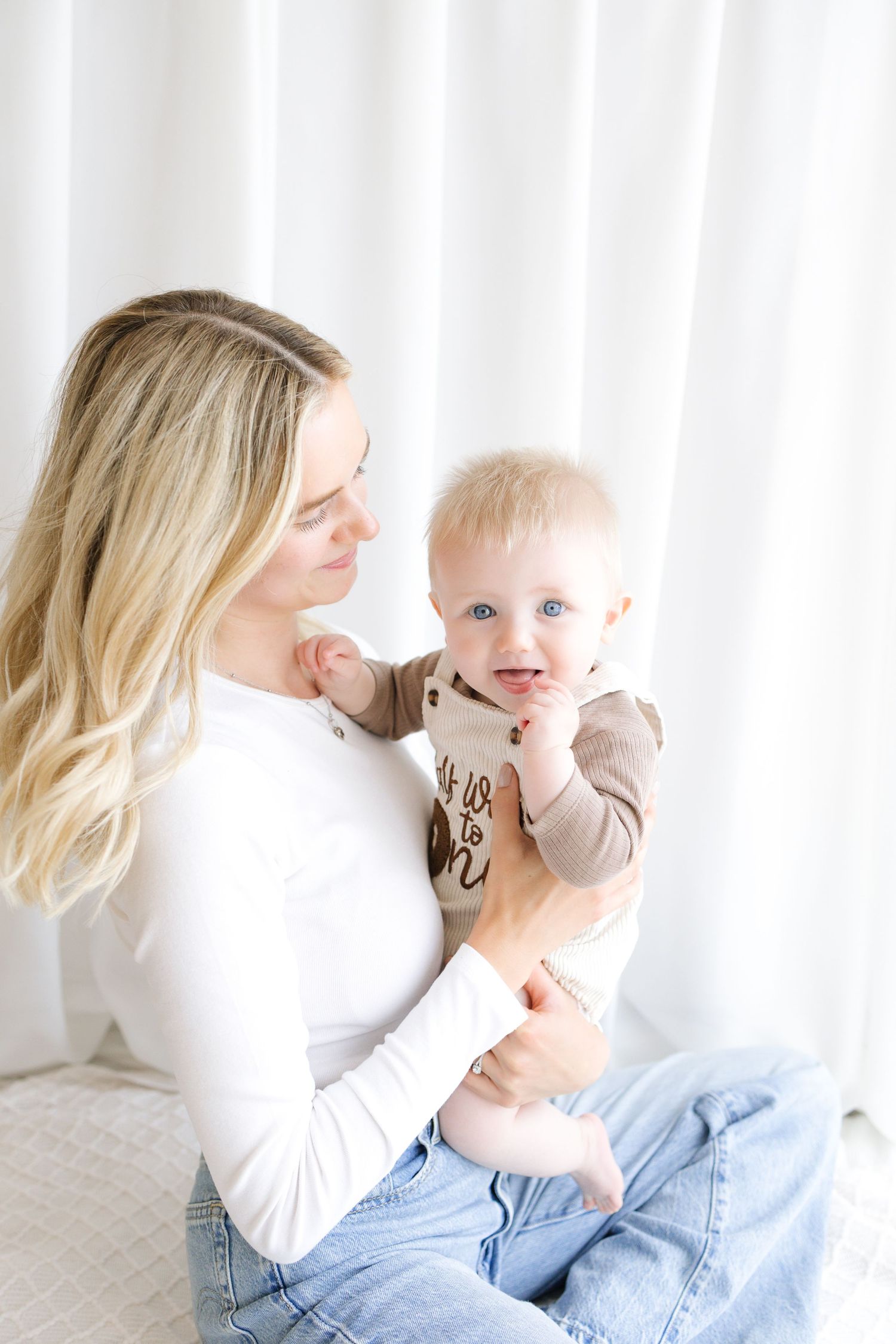 A young blonde mother in a white top and jeans holds her blue-eyed baby, smiling lovingly in a bright, airy room.