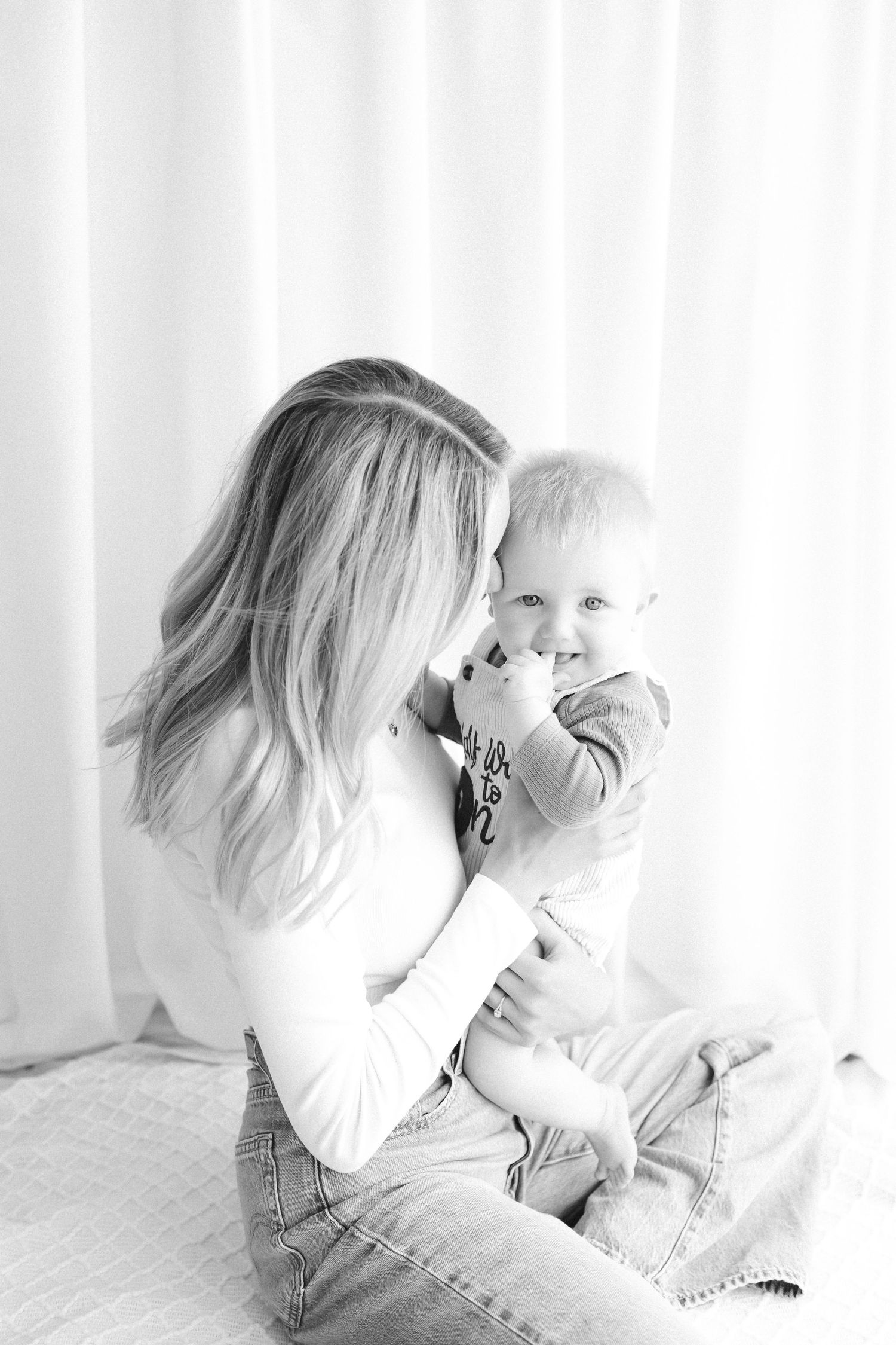Black and white photo of a mother holding and kissing her smiling baby boy while sitting near white curtains.