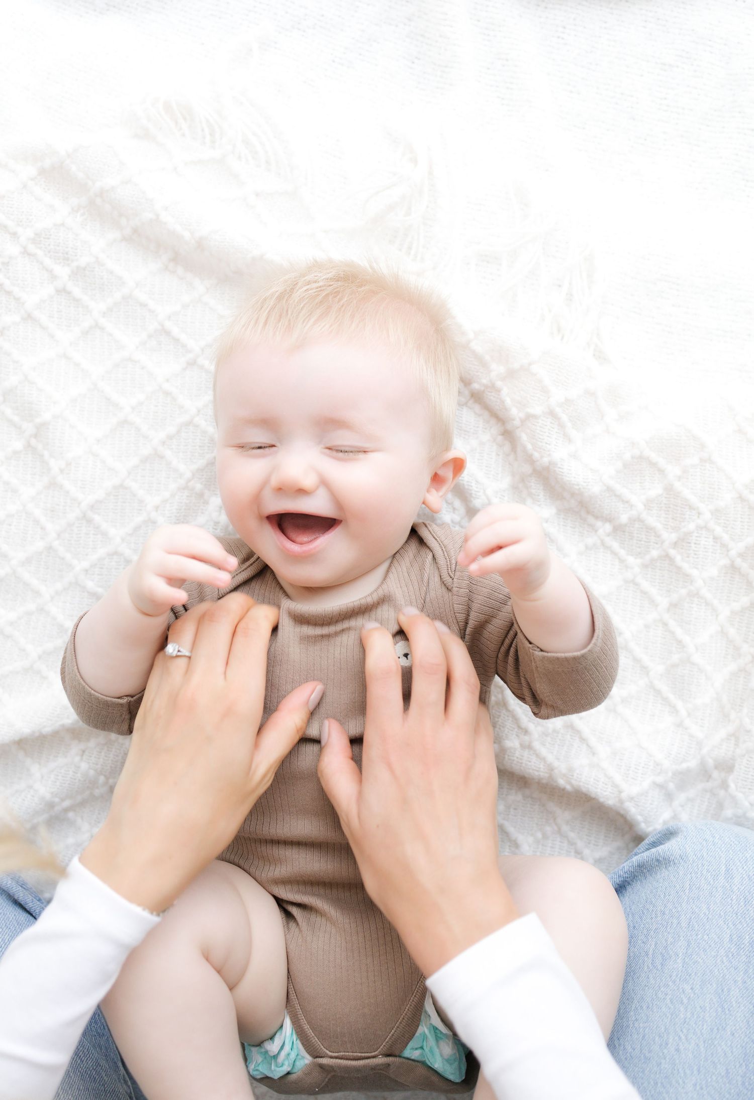 A laughing baby in a brown onesie lies on a white blanket while an adult playfully tickles them.