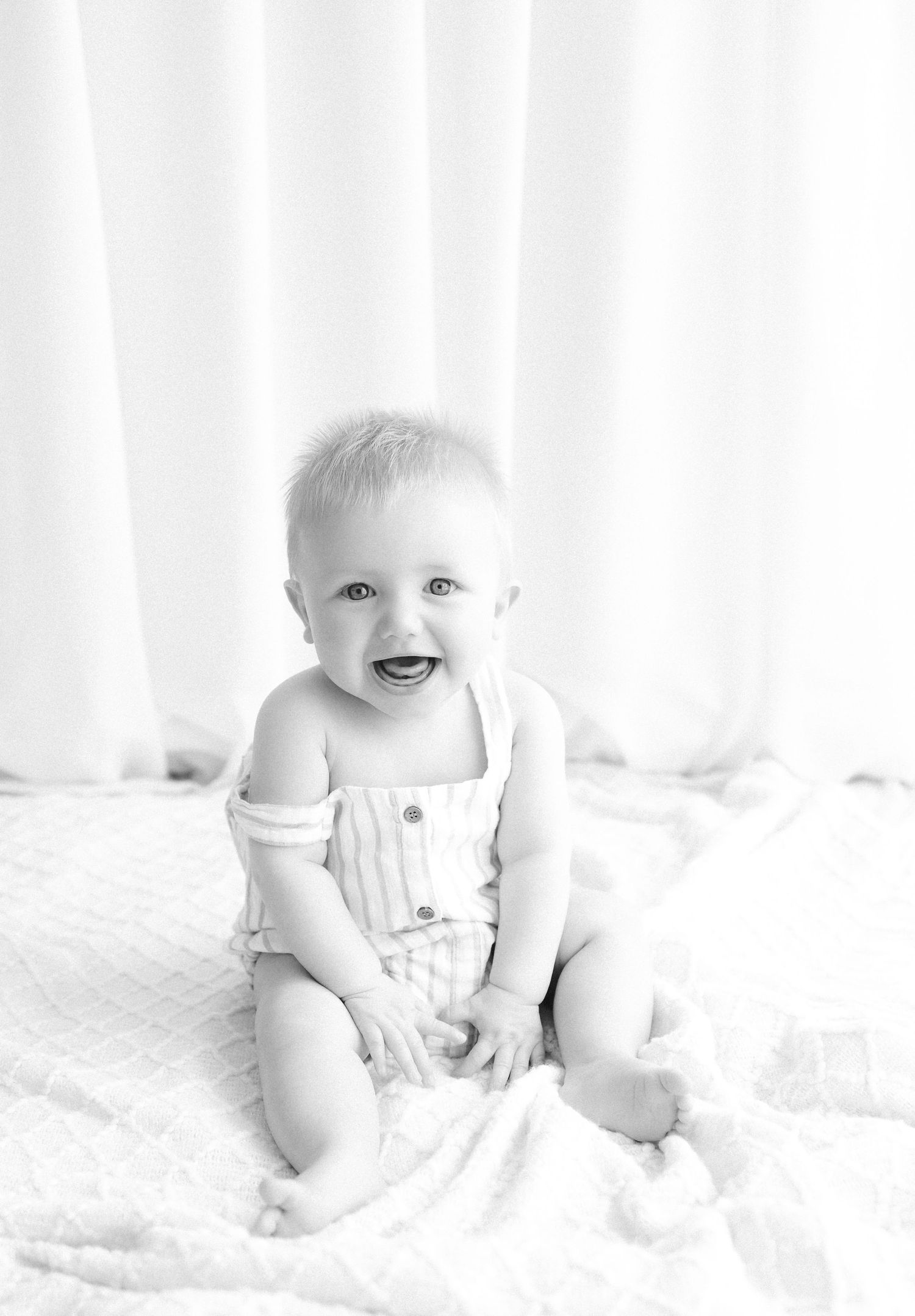 Black and white photo of a happy smiling baby sitting upright wearing a white romper on a soft blanket.