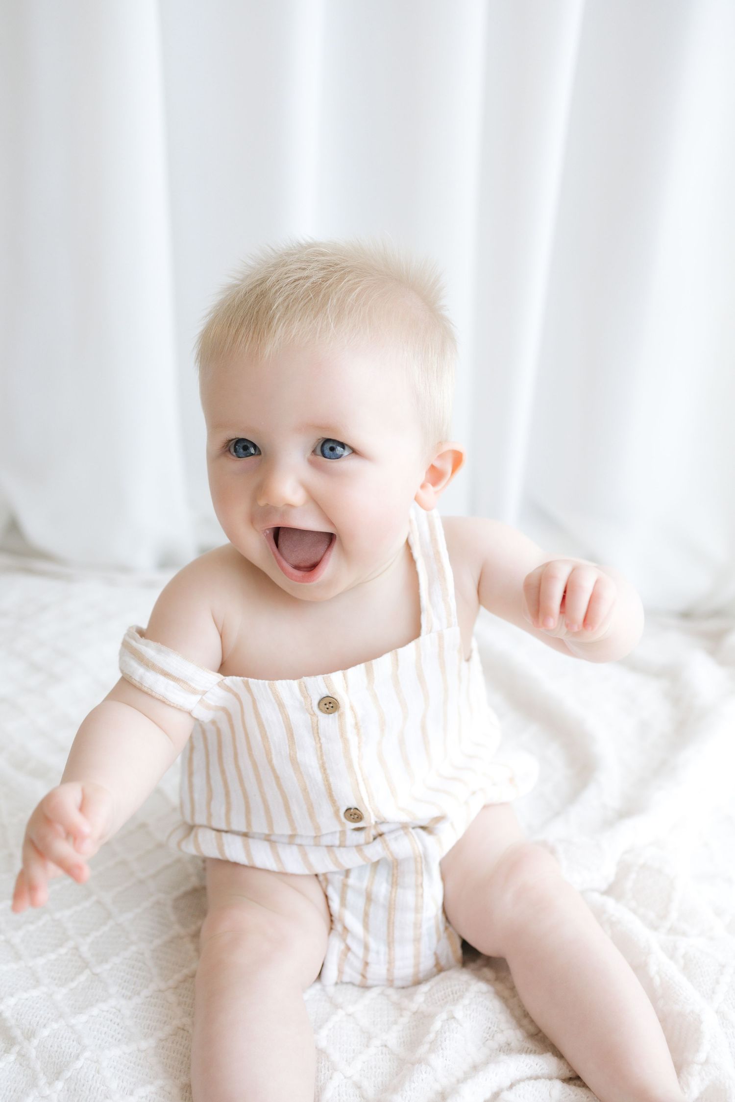 Happy baby with blue eyes wearing striped romper sitting on white blanket, smiling joyfully.