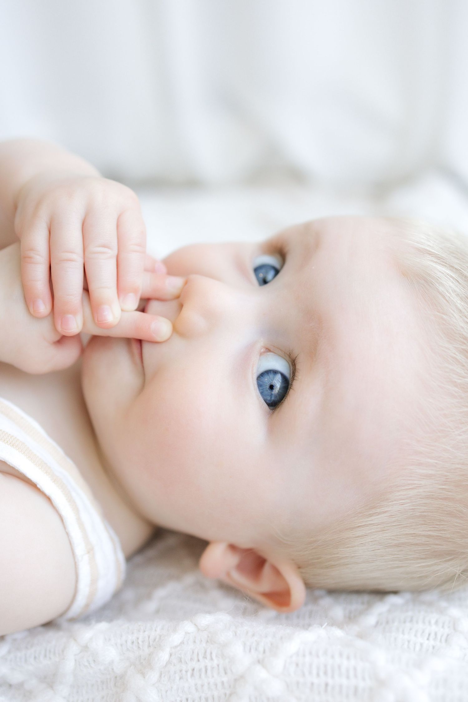 Close-up of a blue-eyed baby lying on a white knit blanket, sucking fingers and gazing upward.