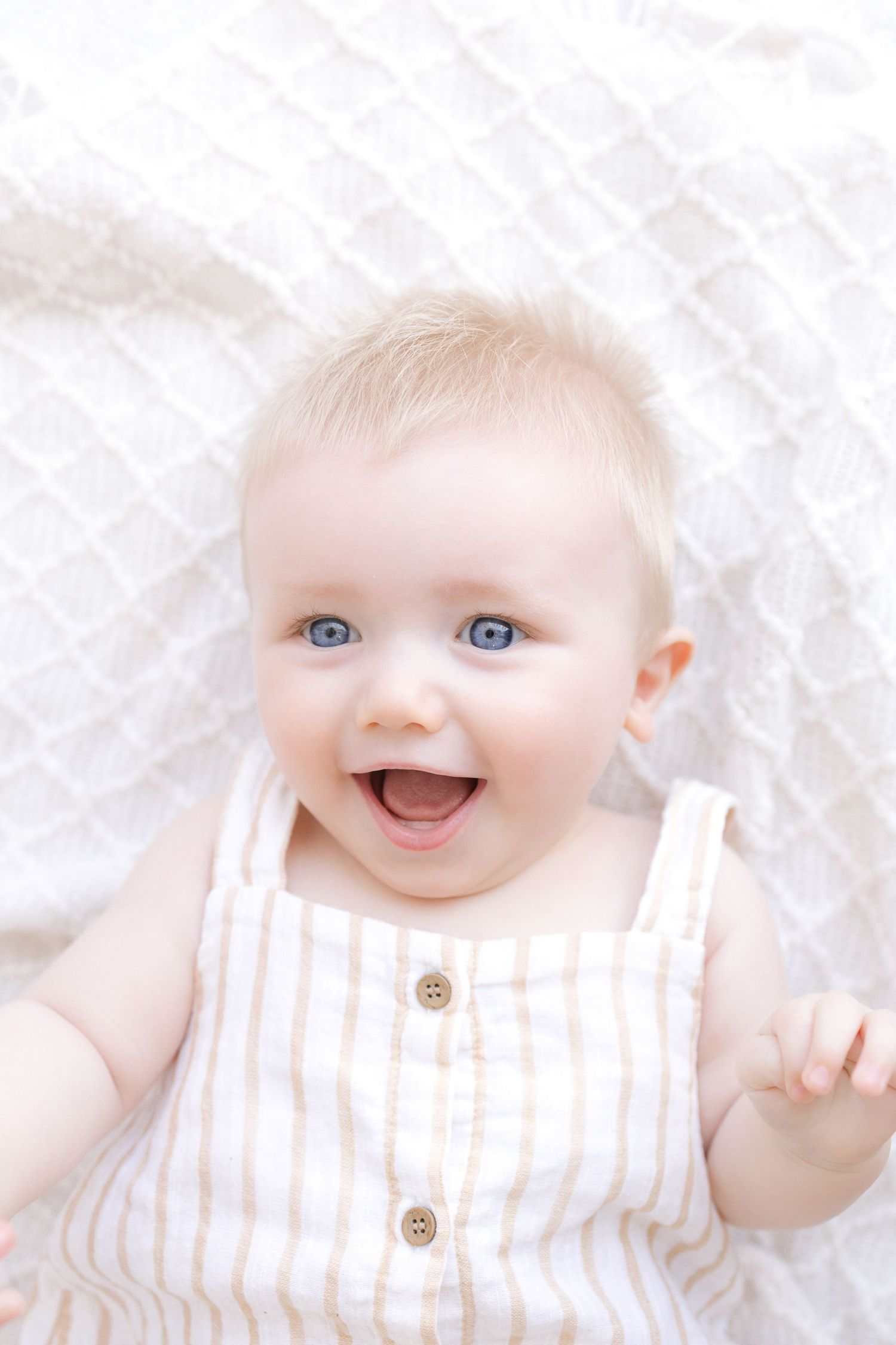 A happy baby with blue eyes and blonde hair smiles while lying on a white blanket wearing a striped sundress.