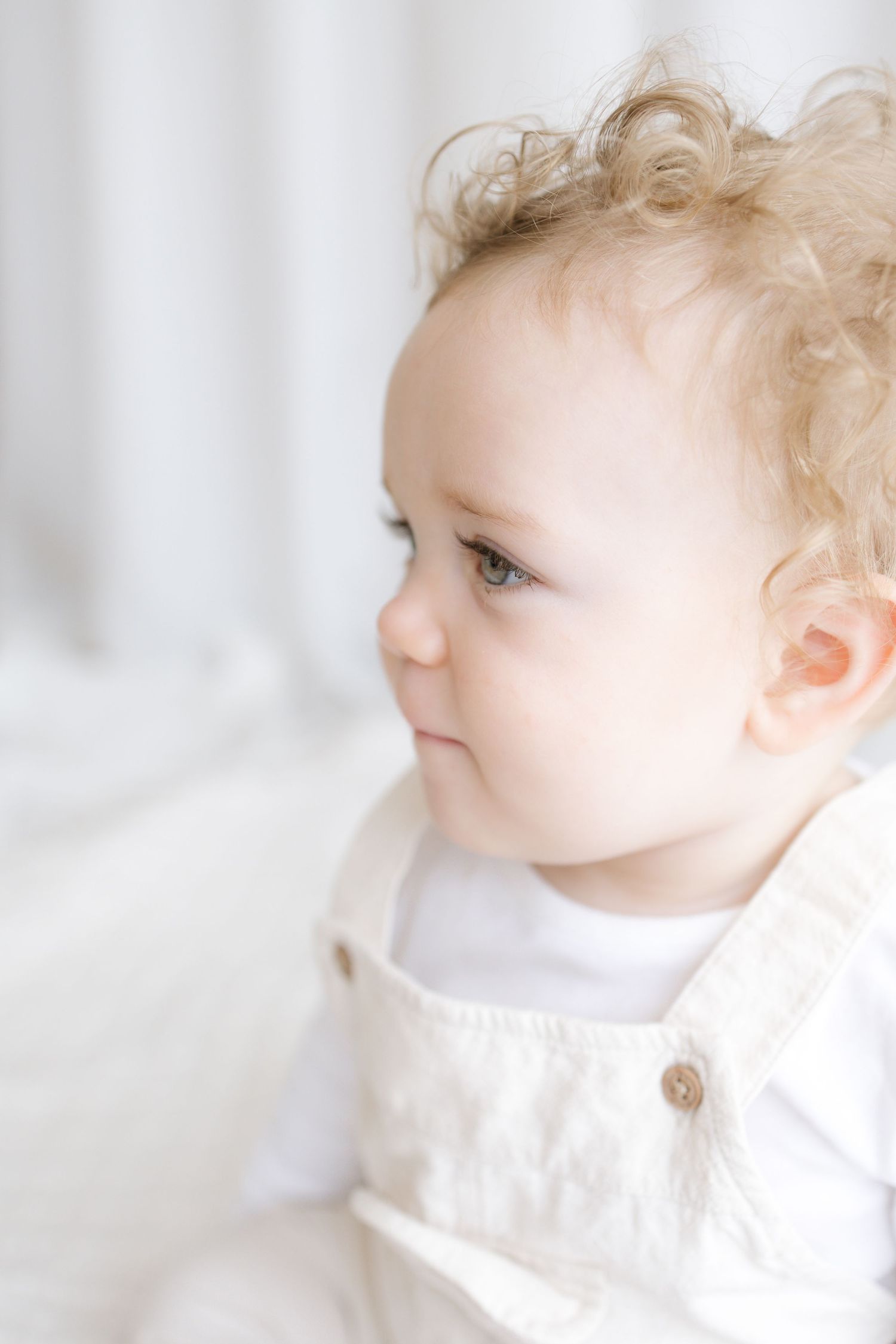 A curly-haired baby in a white linen outfit gazes sideways with bright eyes in a soft, light-filled setting.