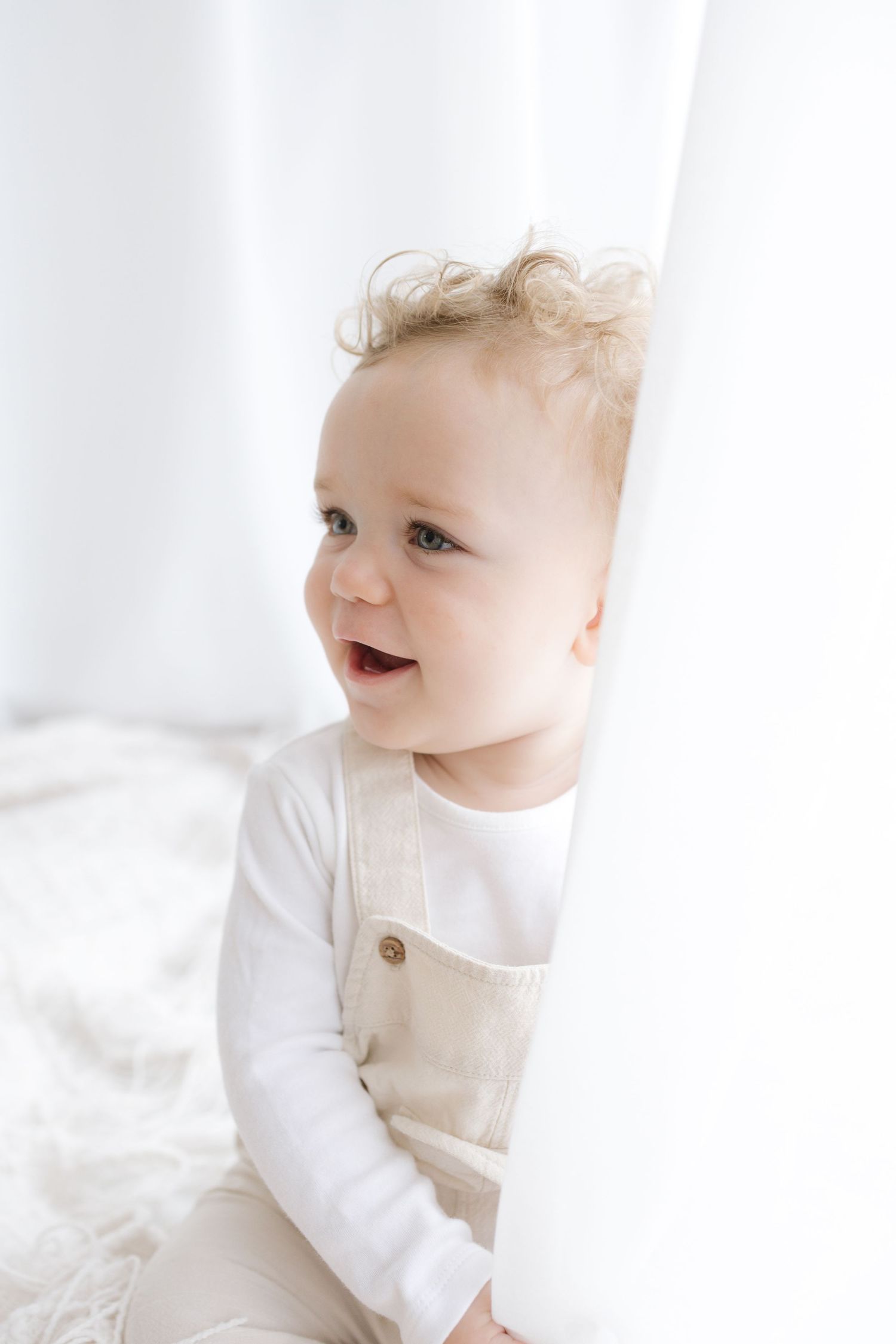 Adorable baby with curly blonde hair wearing a white onesie and beige linen overalls sitting near a white curtain.