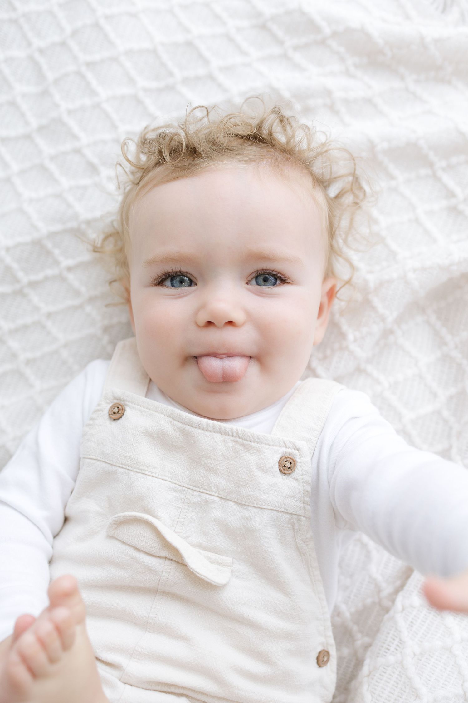 Adorable curly-haired baby in white overalls lying on a blanket, sticking out tongue with a playful expression.