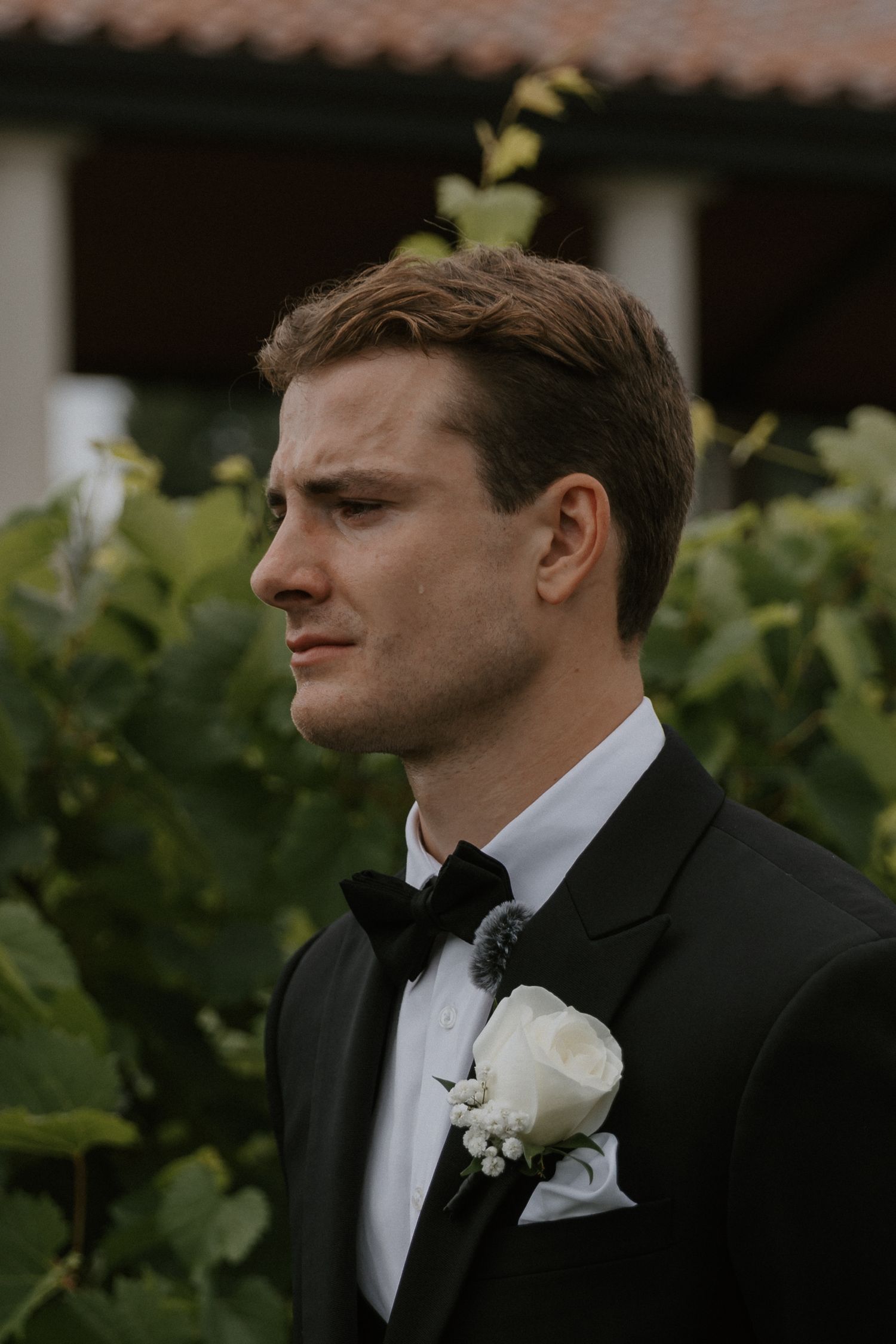 Groom in black tuxedo with white boutonniere against Italian vineyard background.