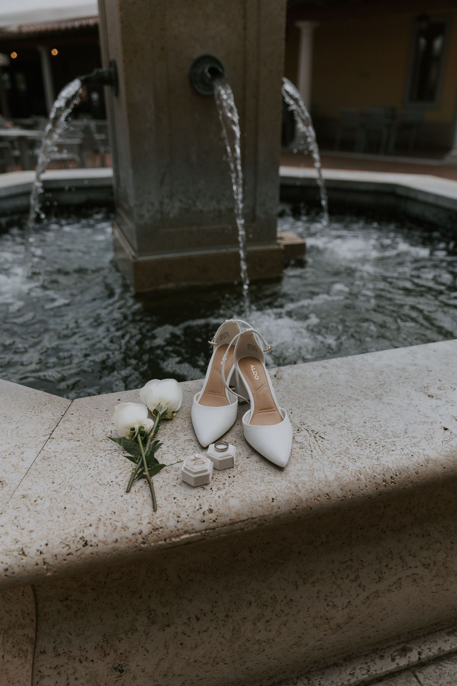 White bridal shoes and wedding rings displayed near an elegant fountain with flowing water in an luxury European-esque outdoor setting.