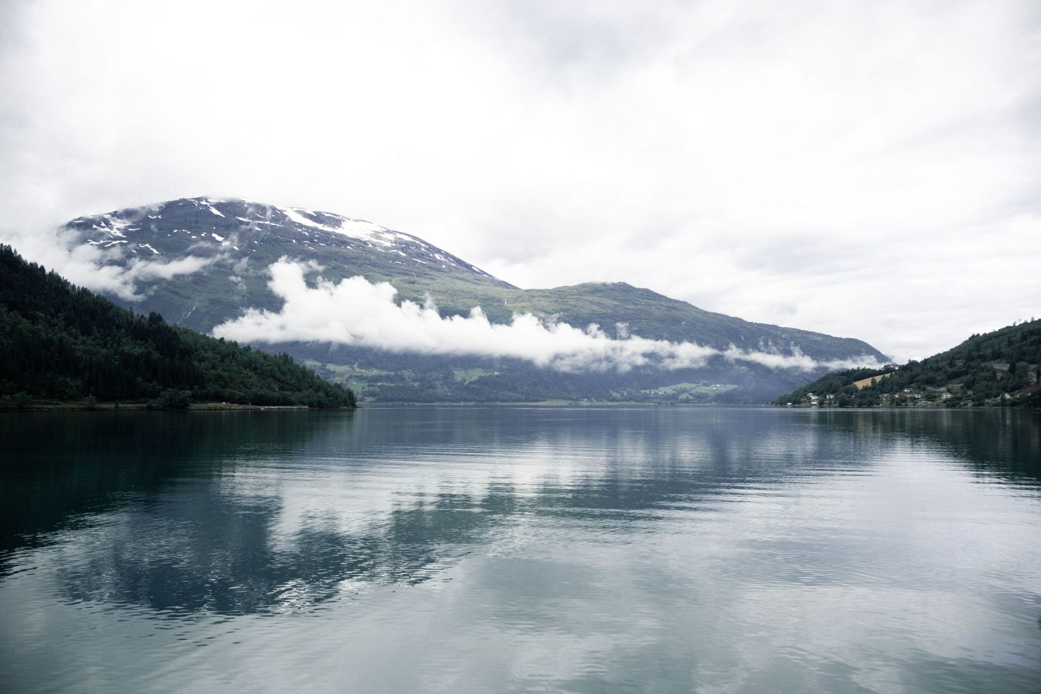 Misty mountain peaks reflect in calm fjord waters under cloudy skies in Norway.