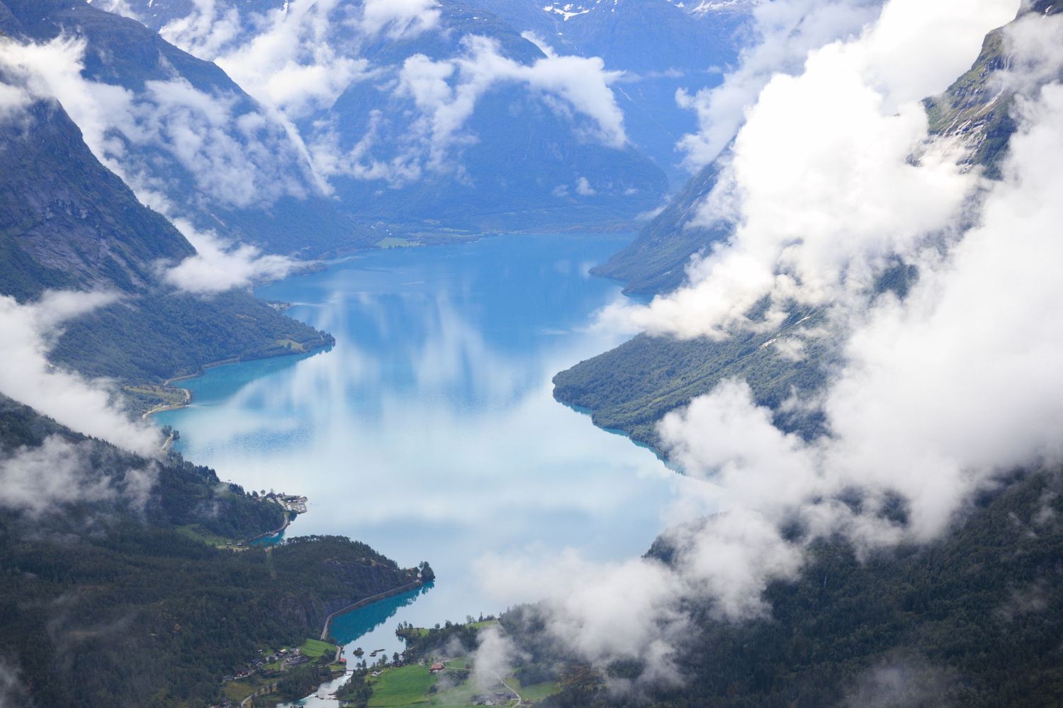 Aerial view of turquoise fjord waters surrounded by snow-capped mountains and white clouds.