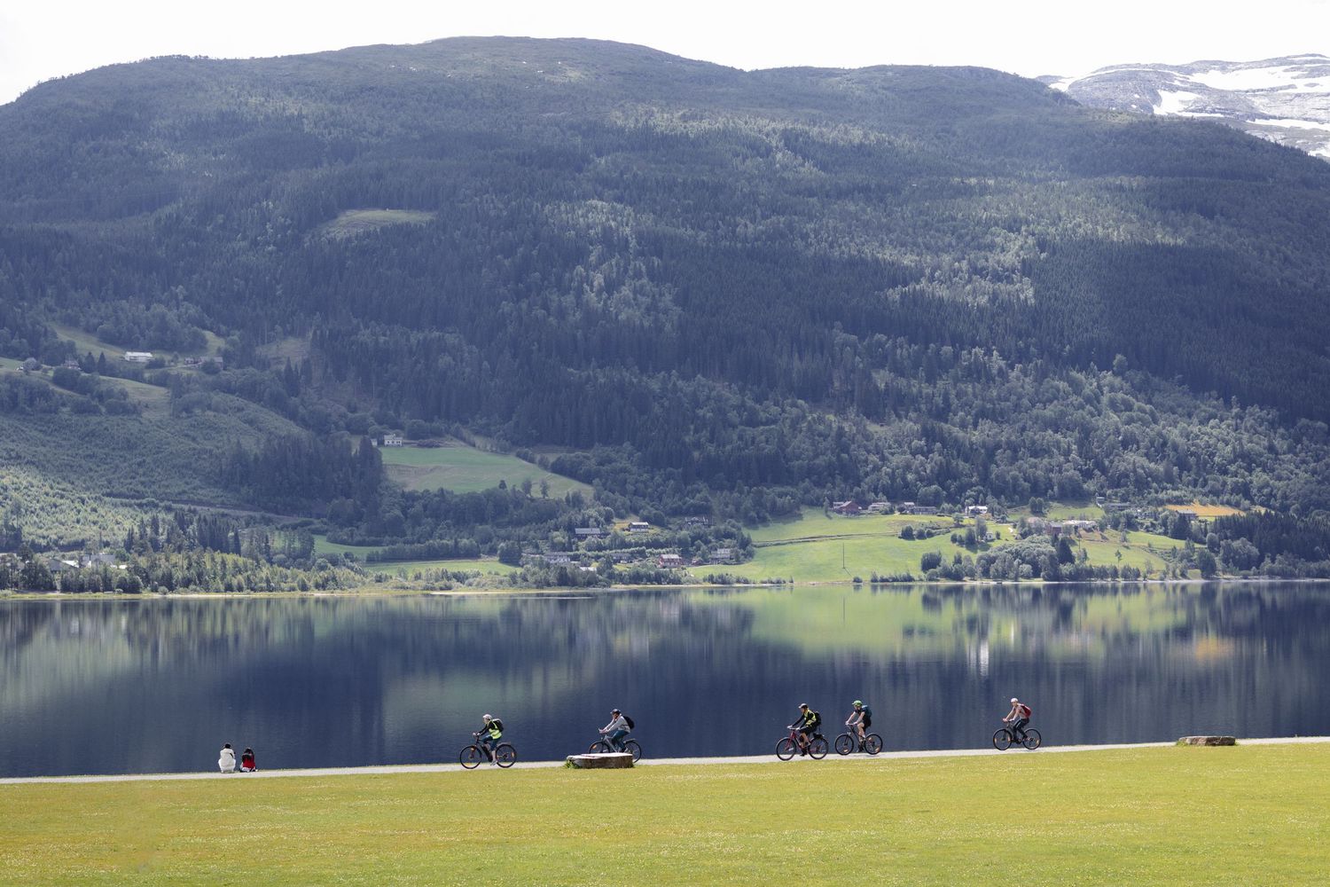 Cyclists riding along a lakeside path with mountains and forest in the background.