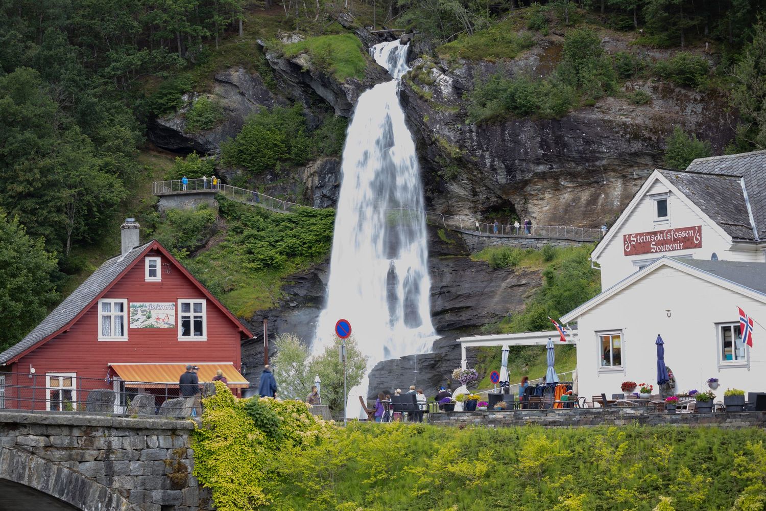 Dramatic waterfall cascades next to red buildings in a Norwegian village with tourists viewing from below.