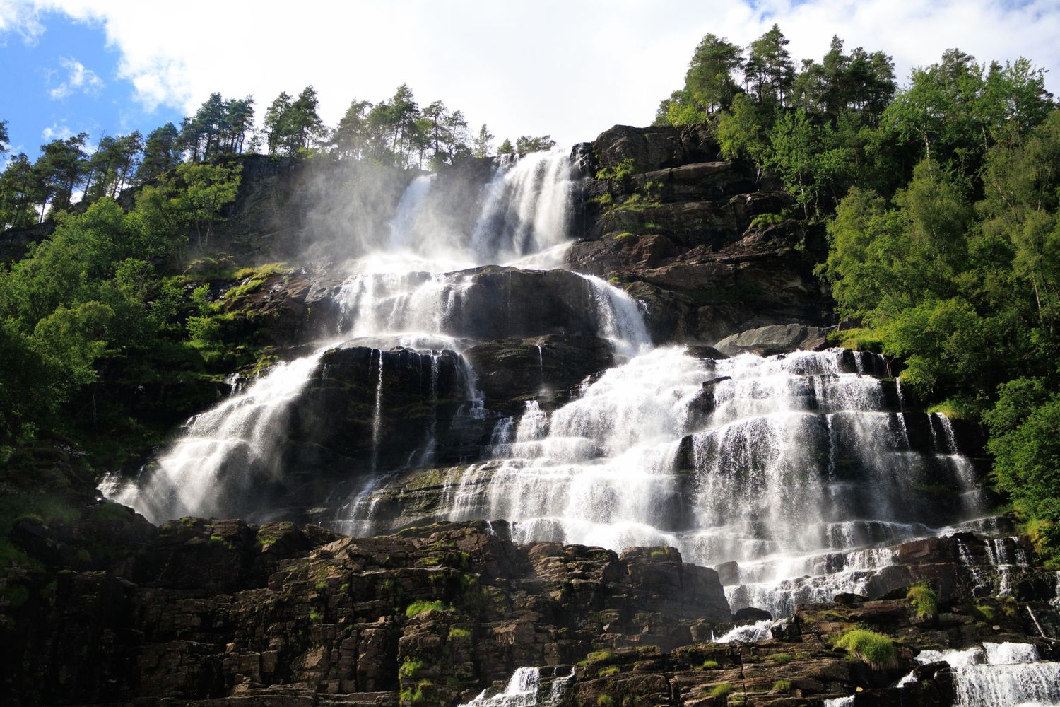 Multi-tiered waterfall flowing down a rocky cliff face surrounded by green forest.