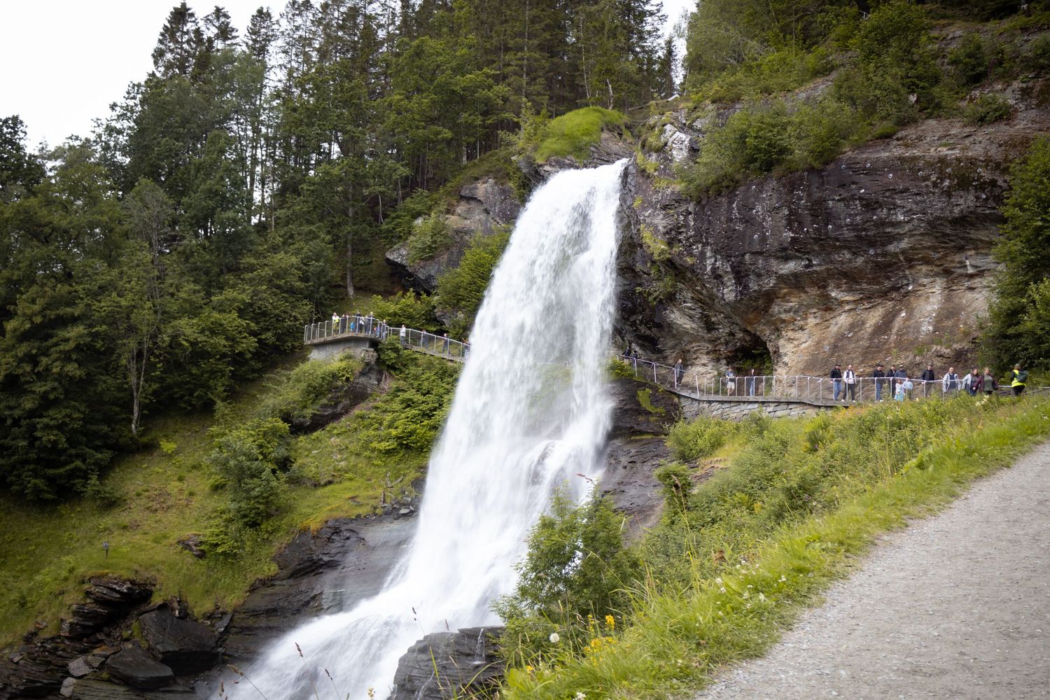 Large waterfall flows down rocky cliff face with hiking path visible in Norwegian landscape.