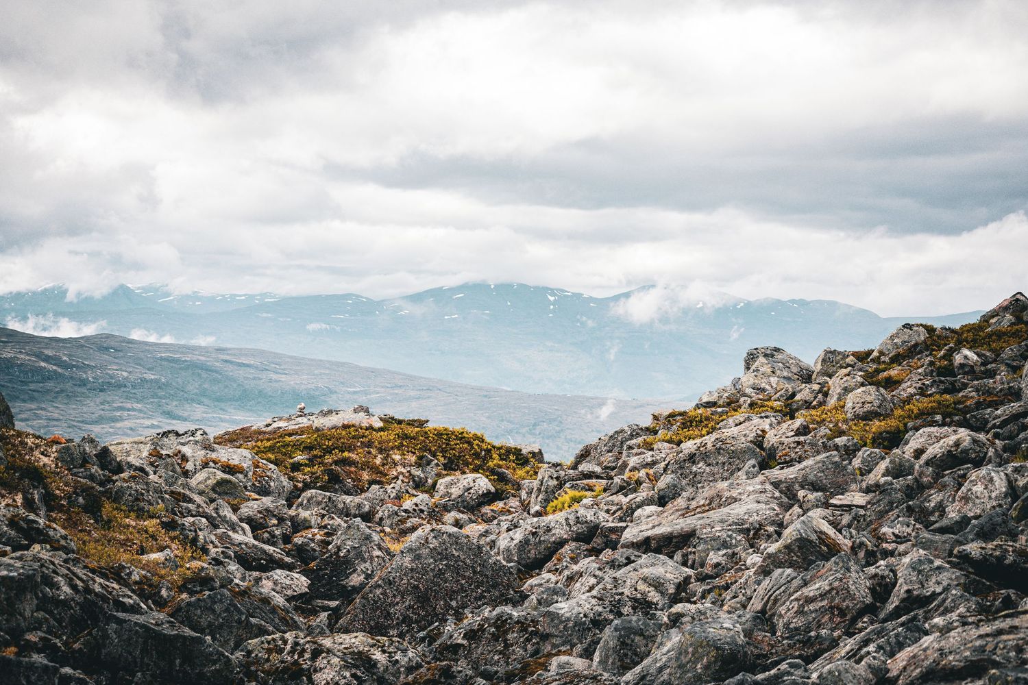 Rocky mountain terrain with cloudy skies and distant mountain ranges in the background.