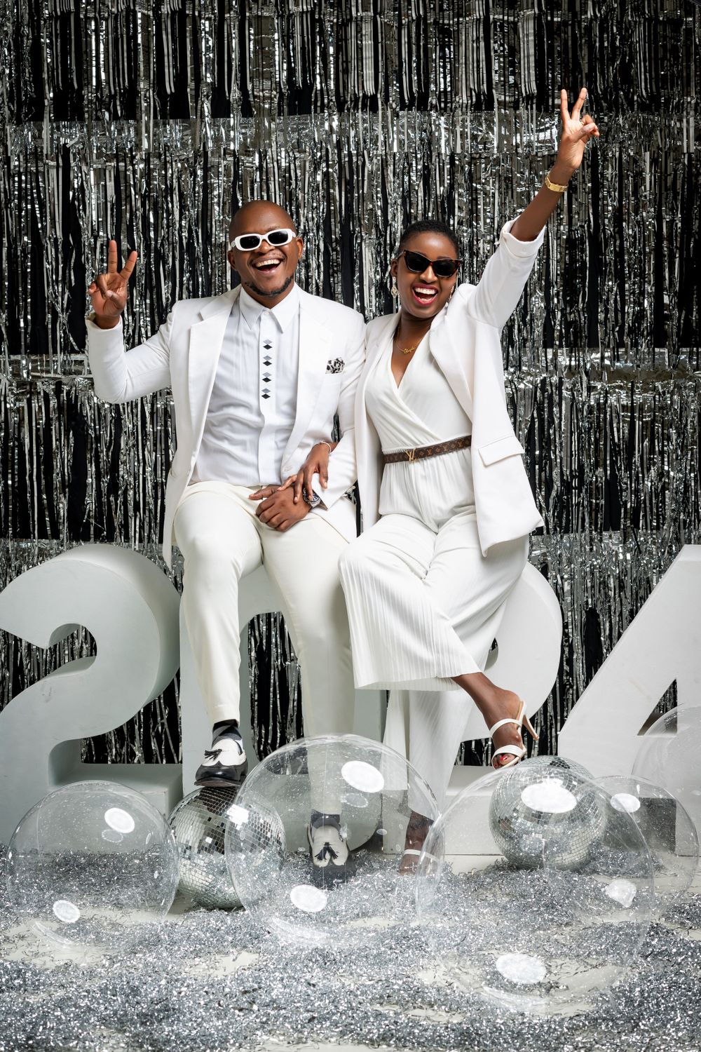 Two people in matching white outfits pose with peace signs against a silver metallic backdrop with 2024 decorations.