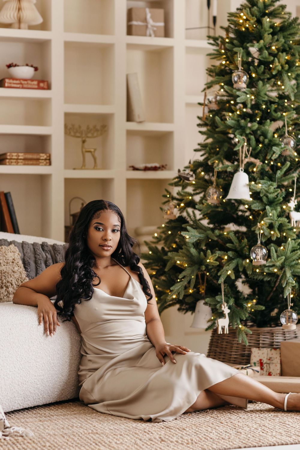 Elegant holiday scene with Christmas tree, built-in shelves, and someone in a cream silk dress relaxing on floor.