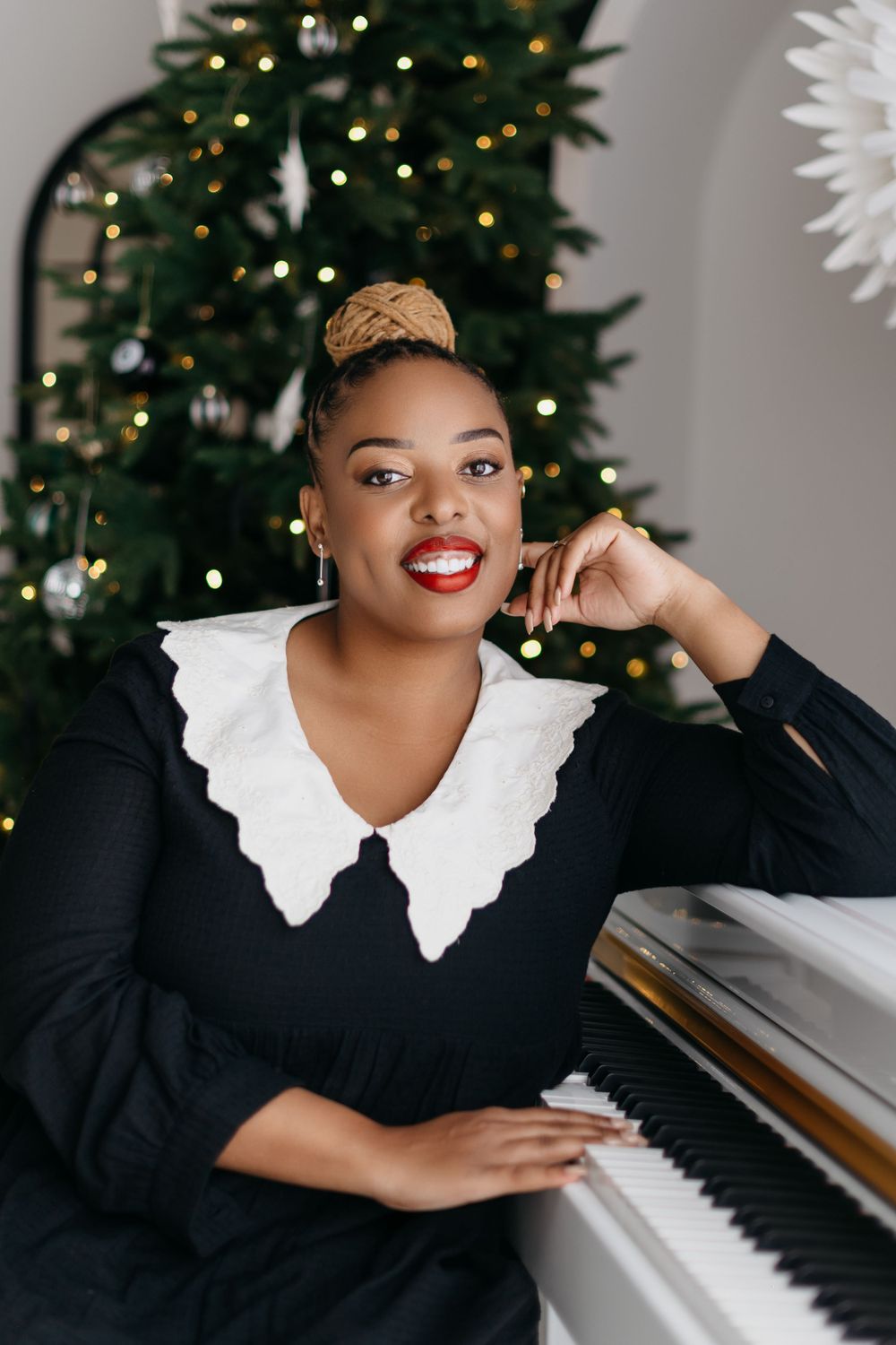 A smiling person in a black dress with white collar poses by a piano with a decorated Christmas tree in the background.