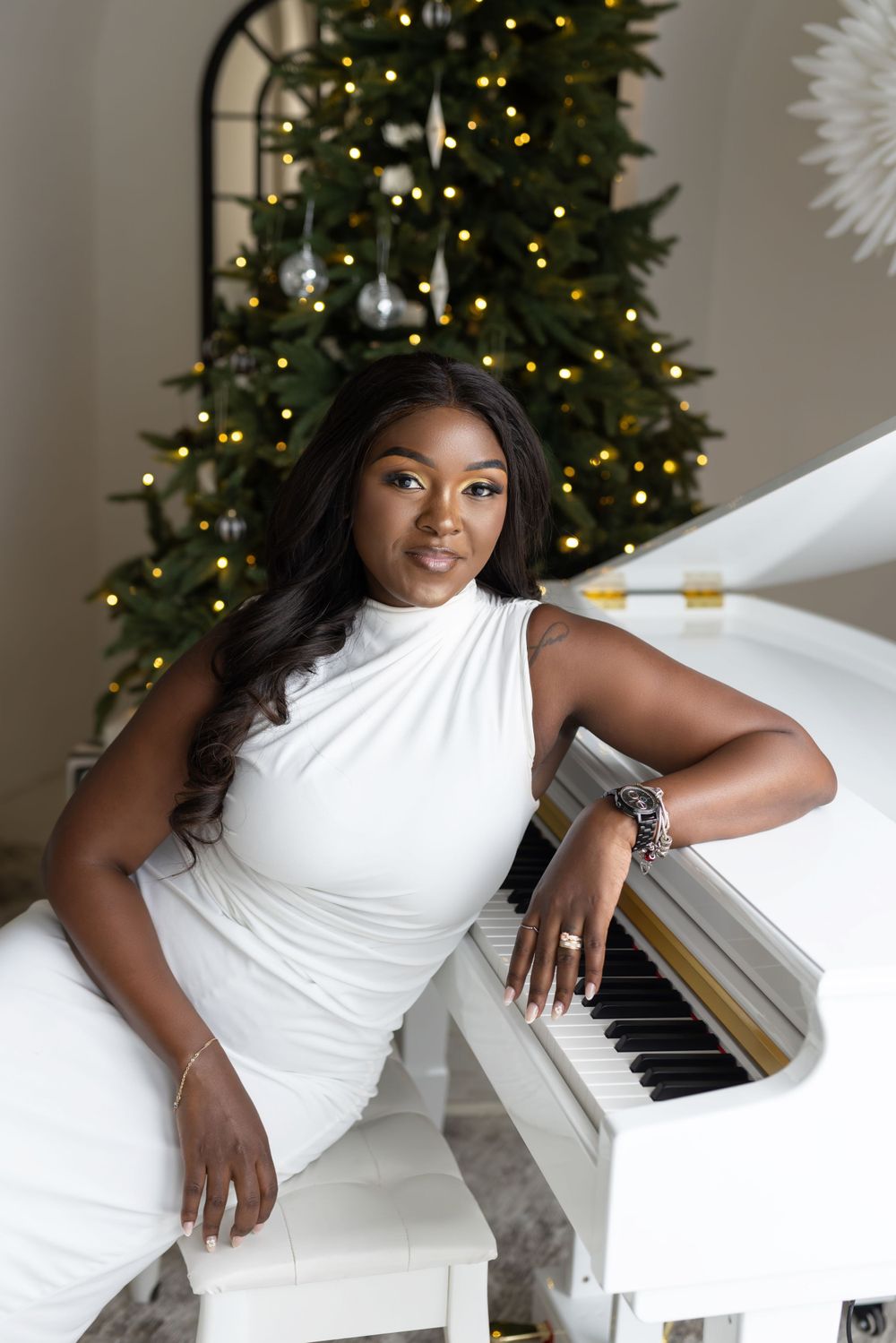 Elegant woman in white dress poses at white piano with decorated Christmas tree in background.