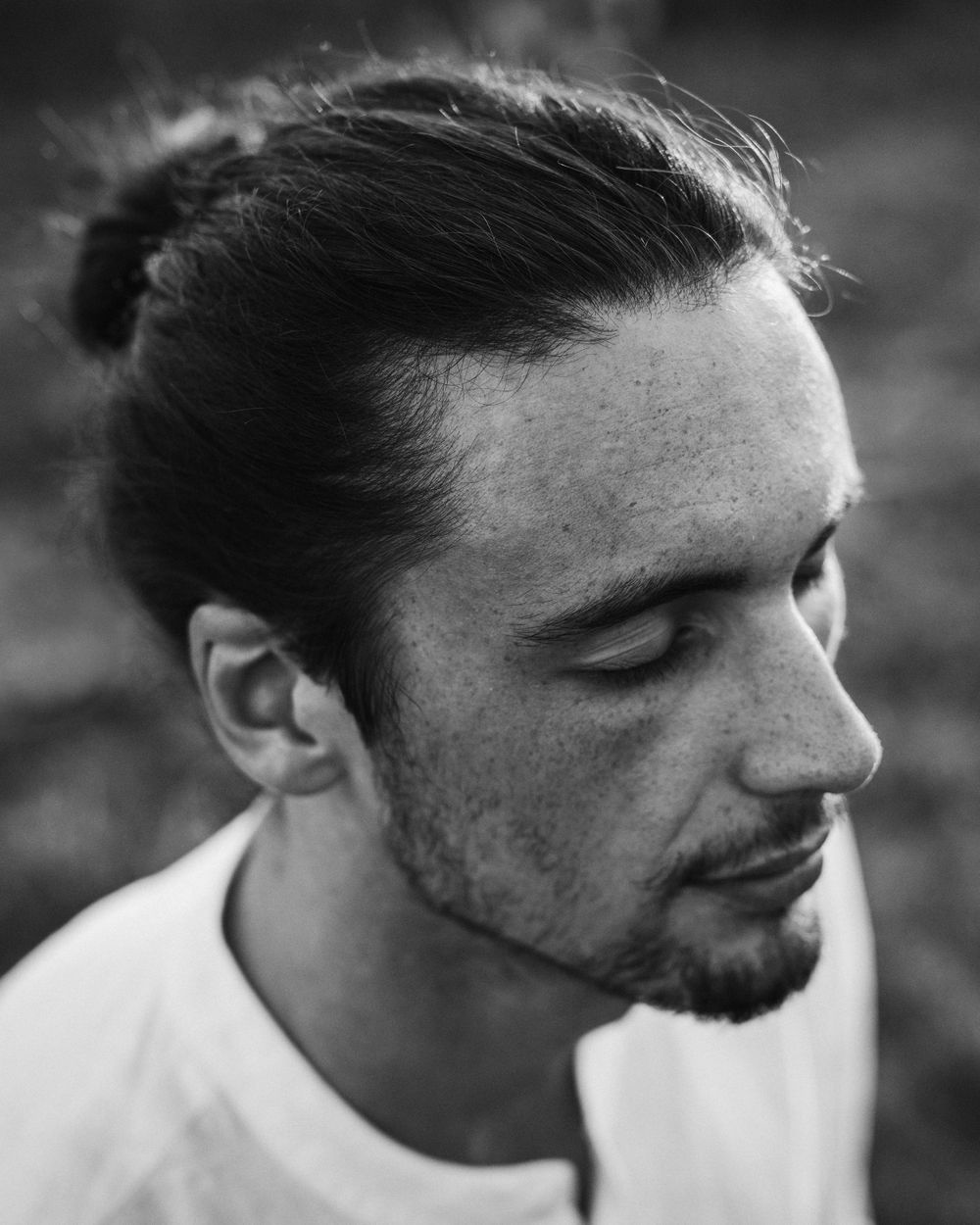 Black and white profile portrait of someone with long hair in a bun and facial hair looking down while wearing white.