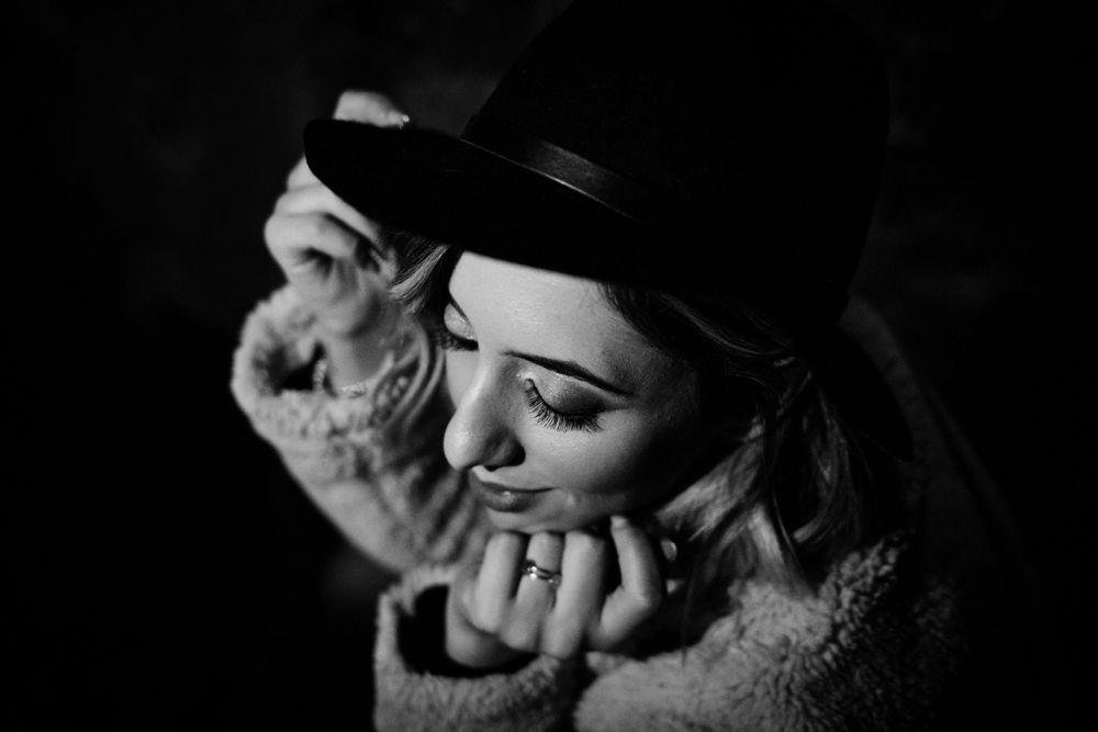 Black and white portrait of a figure in shadow wearing a hat and gently resting their chin on their hand and jacket collar.