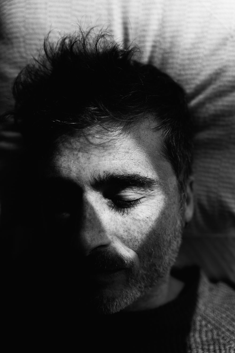 A dramatic black and white close-up portrait of a person with facial hair resting with eyes closed on a pillow.