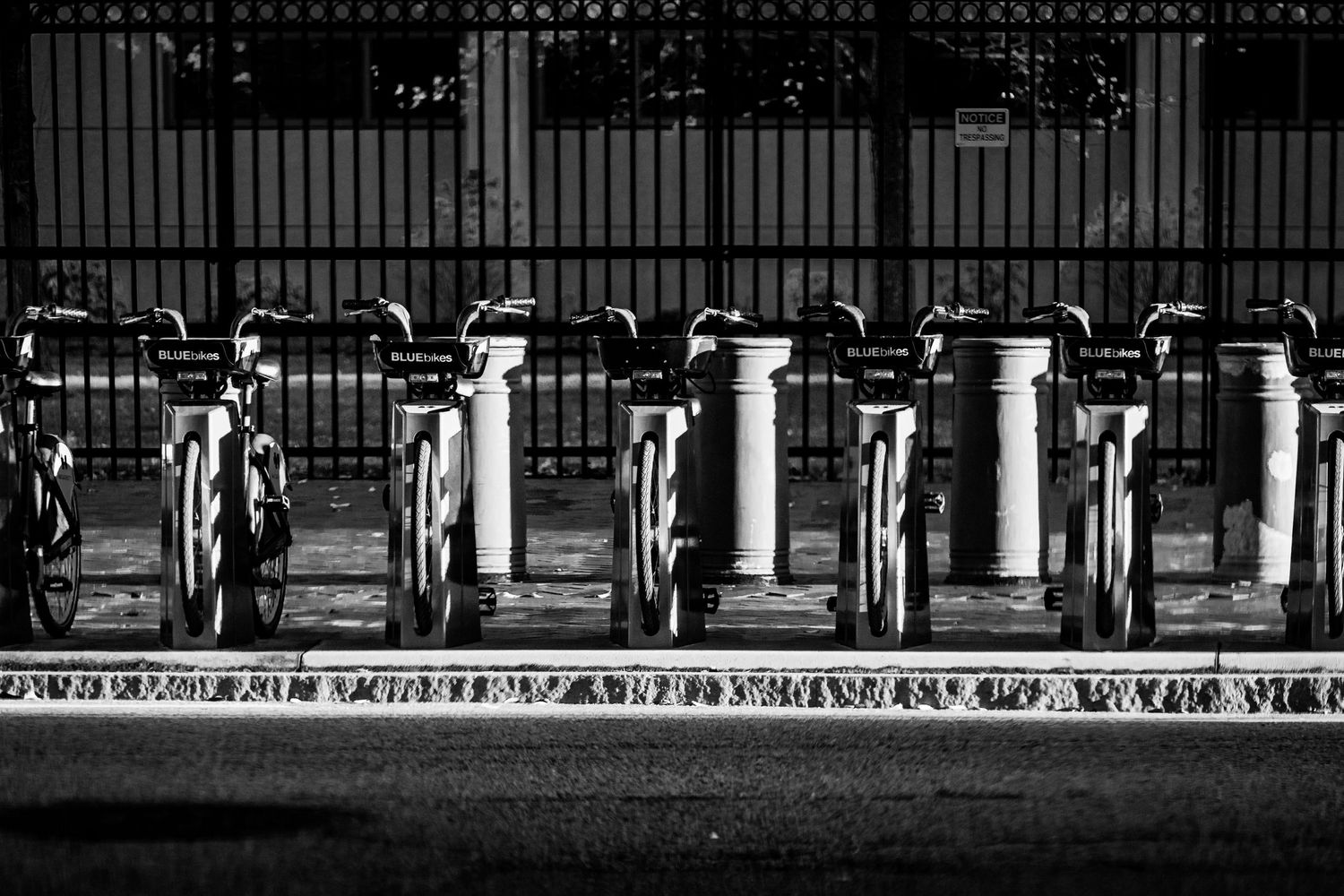 A black and white photograph shows a row of vintage gas pumps behind an iron fence.