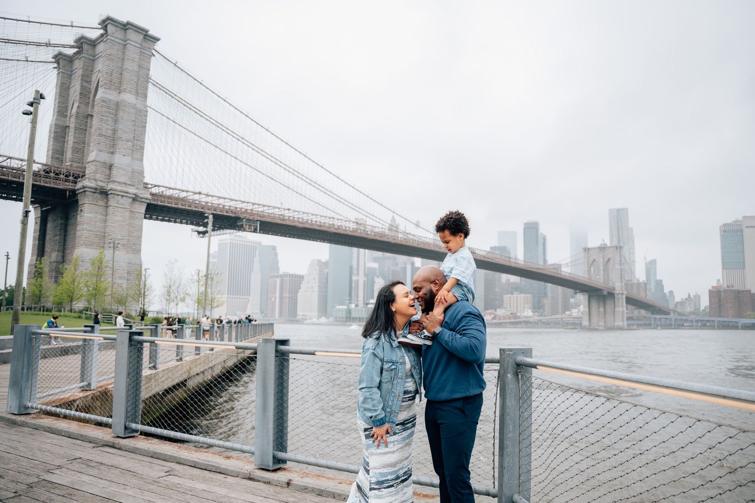 Couple embraces on waterfront promenade with Brooklyn Bridge and Manhattan skyline in background.