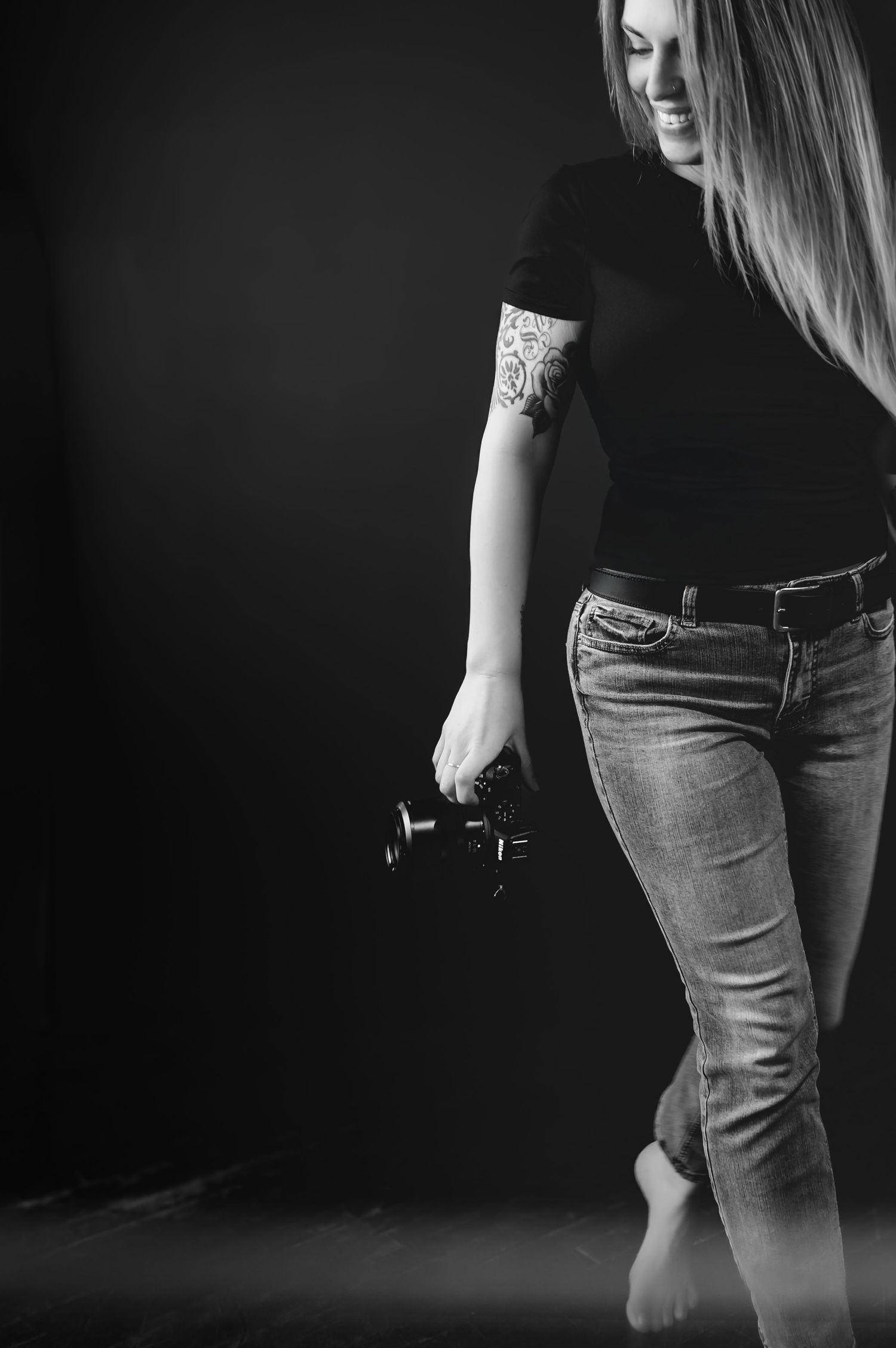 Photographer in plaid shirt and dark pants holds two DSLR cameras while striking playful barefoot pose against tan backdrop.