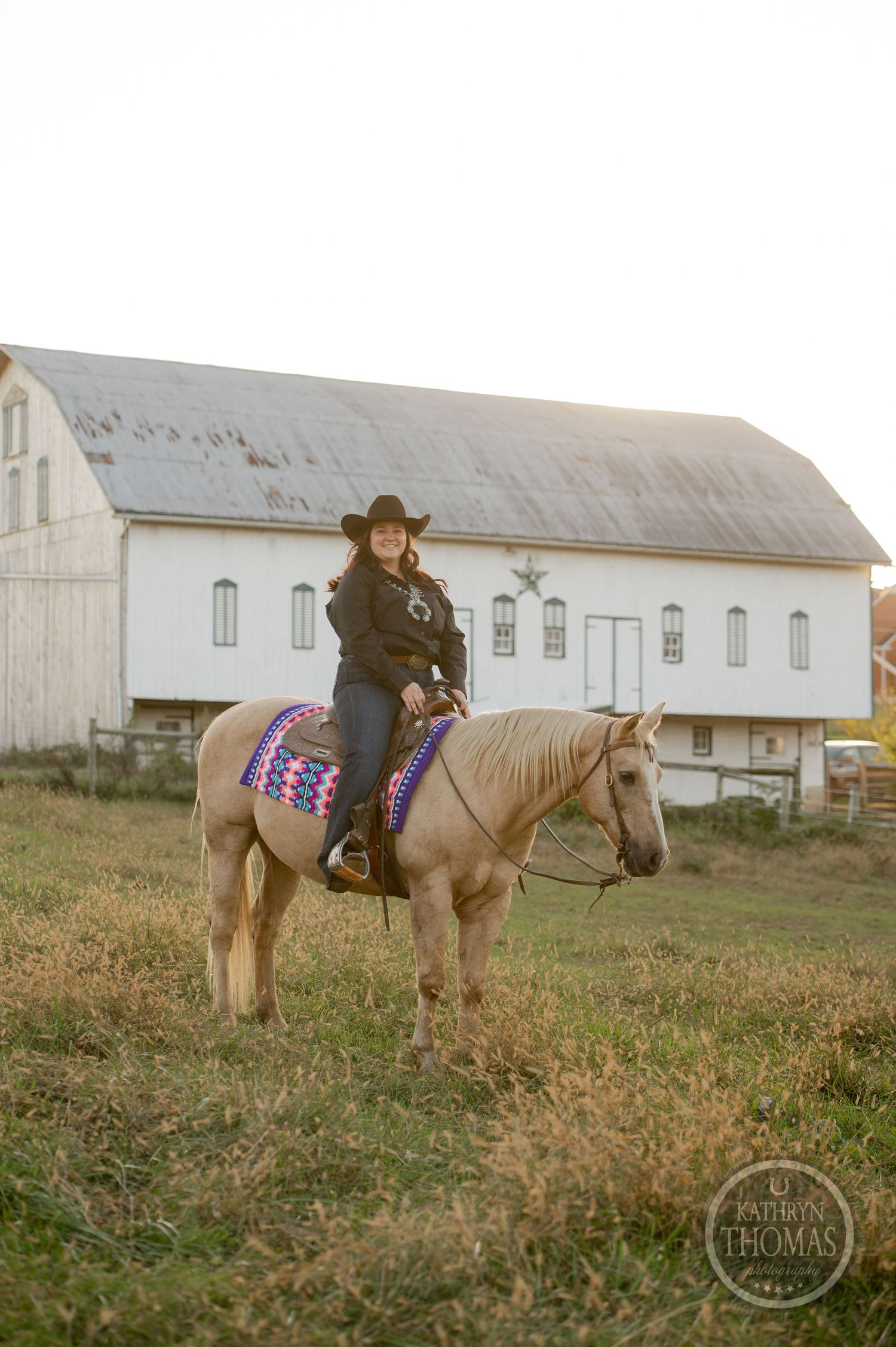Pale horse with western tack and colorful saddle blanket stands by white barn.