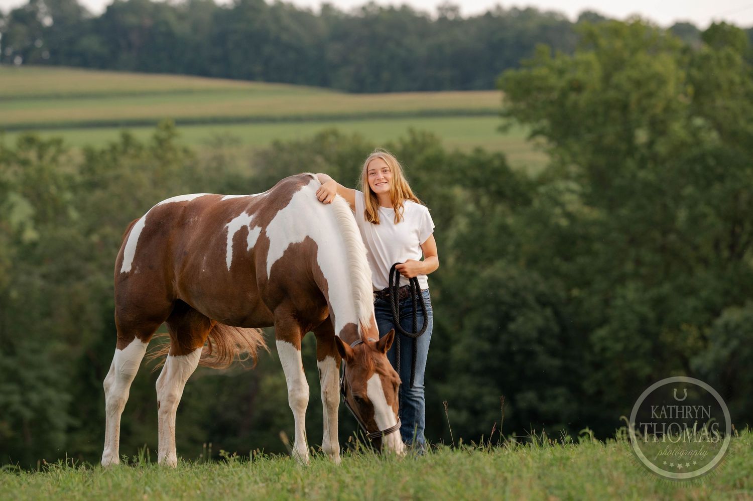 A paint horse grazes while someone stands nearby in an evening pasture.